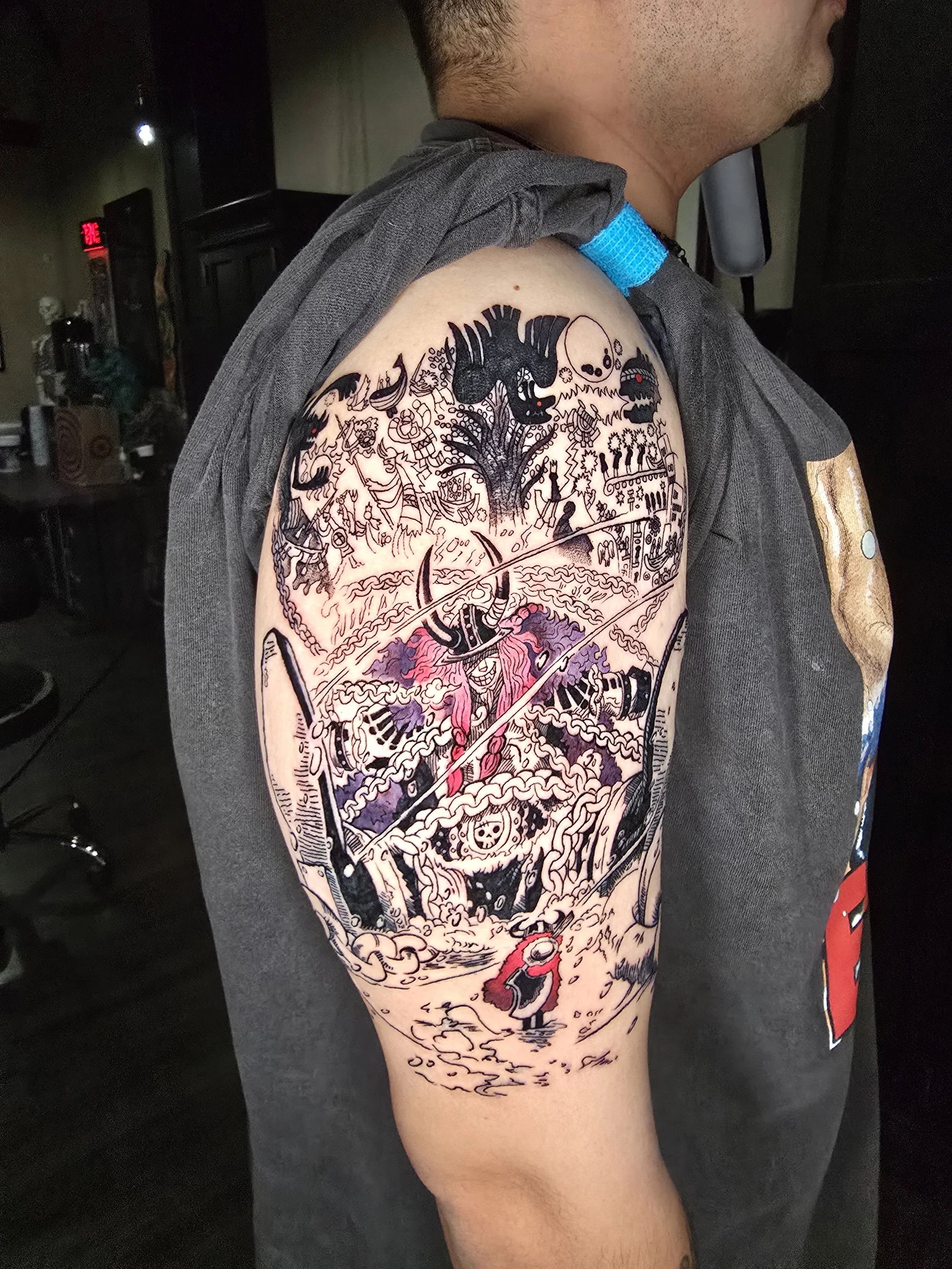 A tattoo on a person's upper arm depicting a stylized mountain scene with various creatures, including a dragon and a skeleton, in black ink with detailed line work and some red accents.