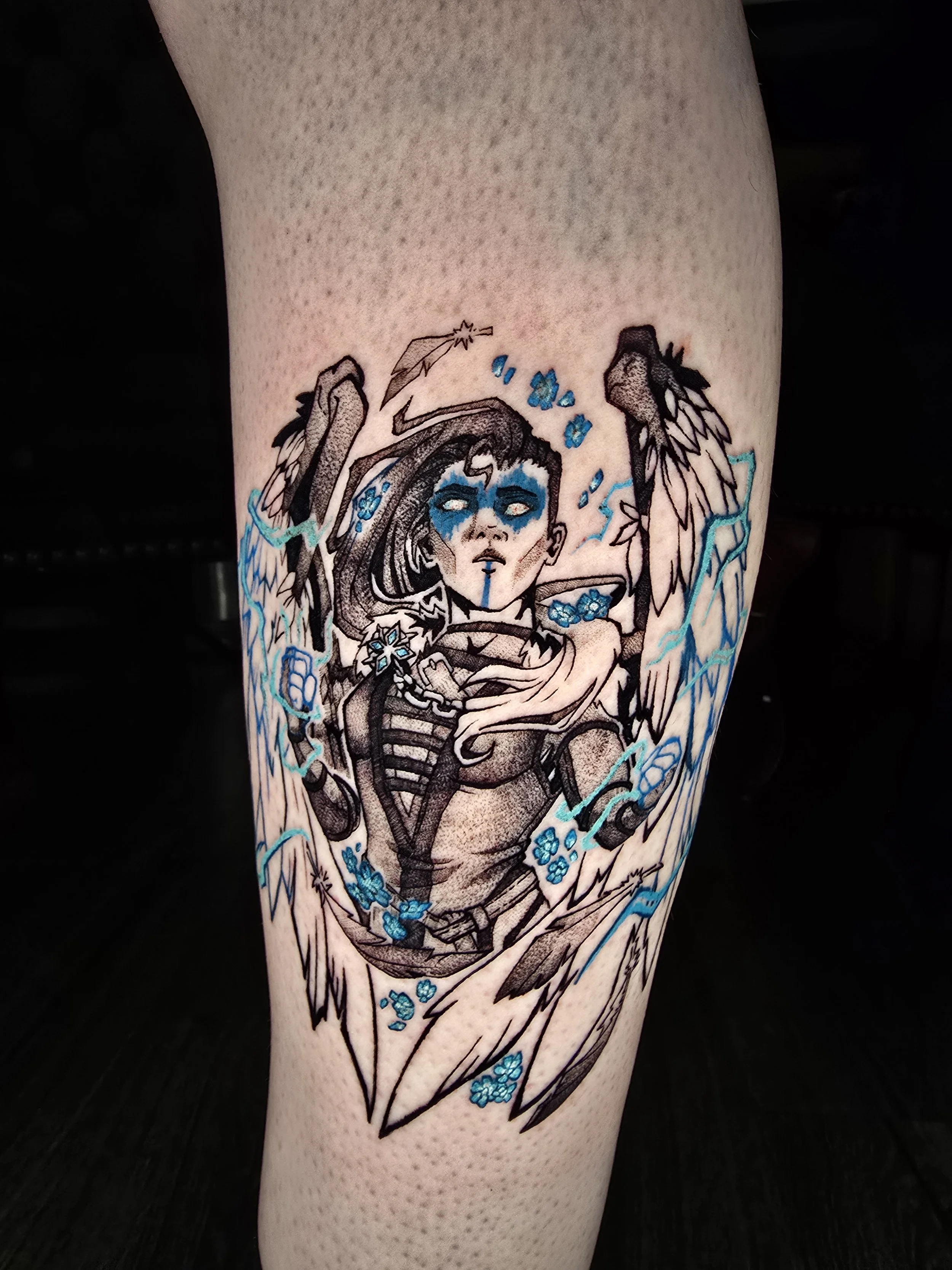 Tattoo of a skeleton with a woman’s head and long hair, surrounded by flowers and lightning, on a person's leg.