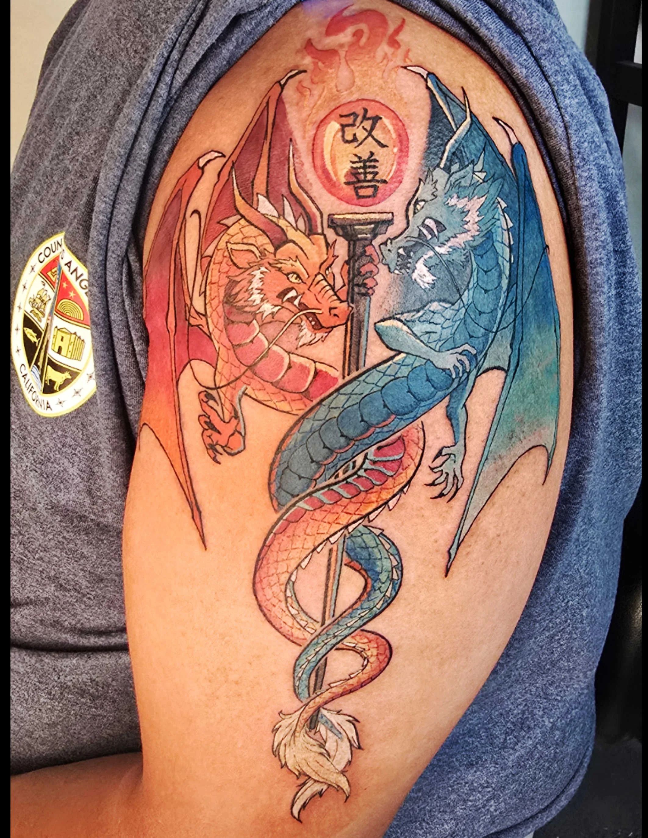 Colorful tattoo of a dragon and a creature with wings, holding a staff with a circular symbol and Chinese characters, on person's upper arm.