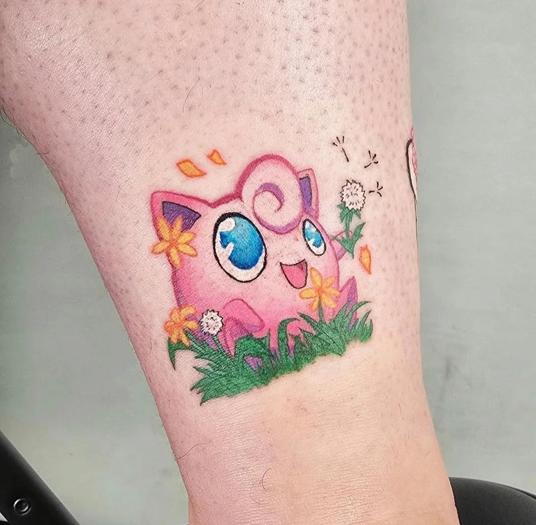 Colorful tattoo of Jigglypuff from Pokémon, surrounded by grass and flowers on a person's skin.