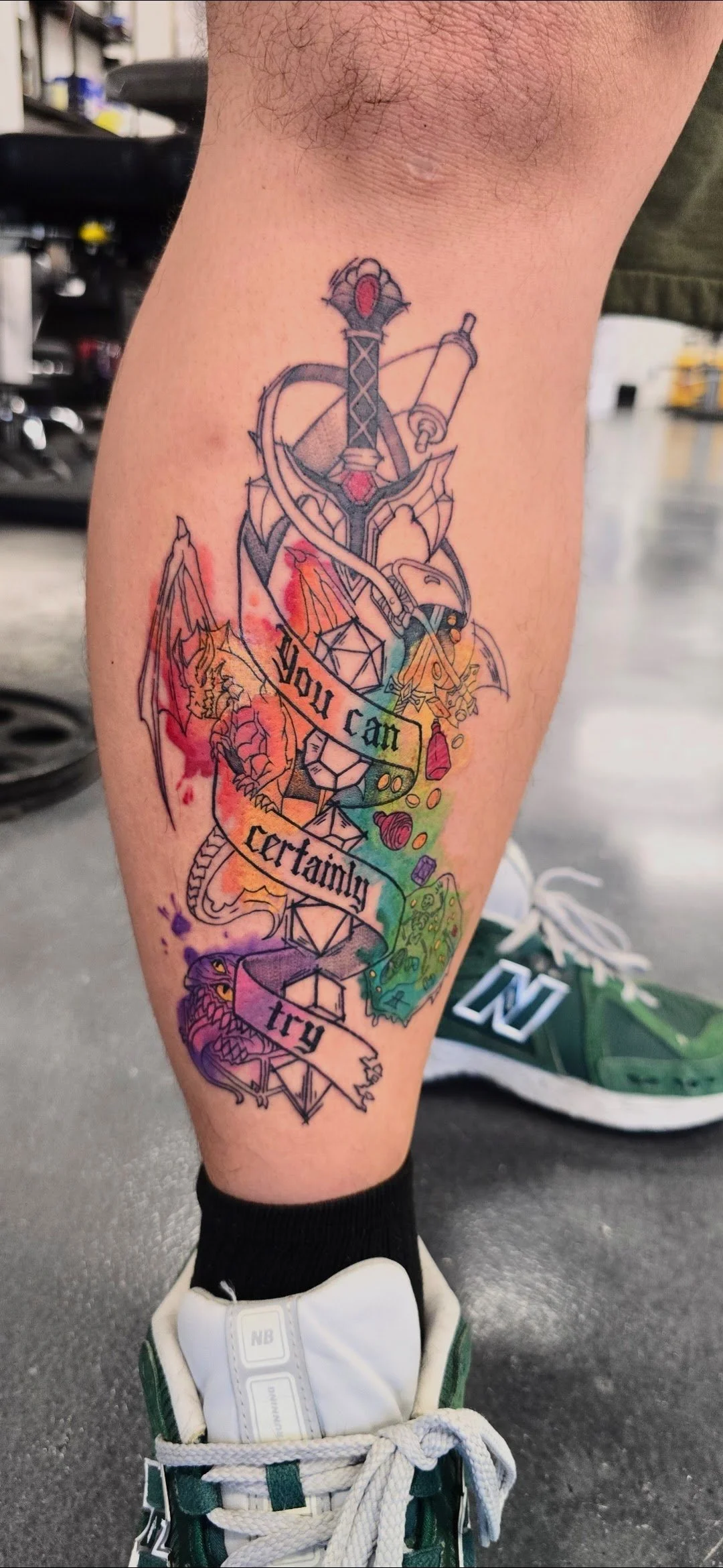 Colorful tattoo on a person's calf featuring a sword, a dragon, a bird, and various fantasy elements, with the phrase 'You can certainly try' written across it.