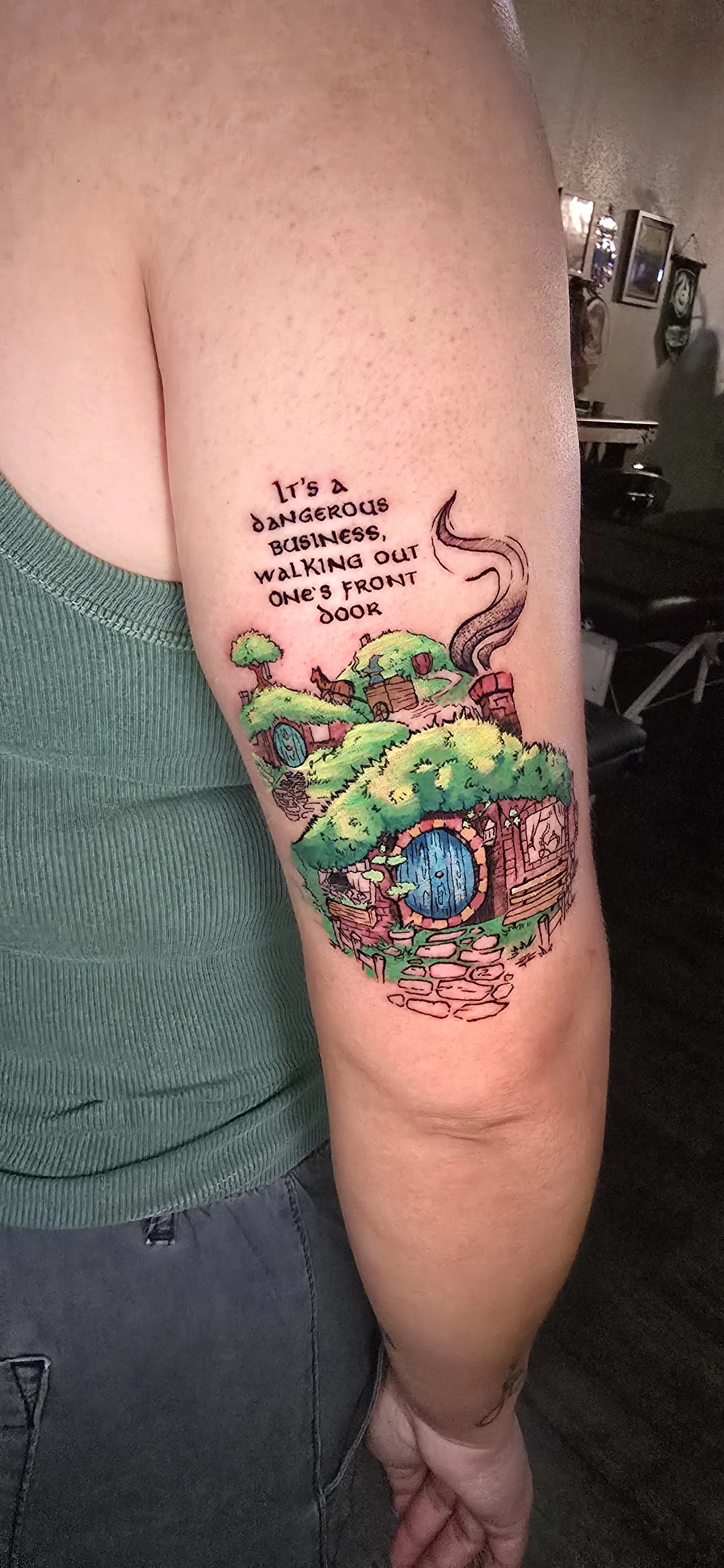 Colorful tattoo on arm depicting a whimsical cottage with a blue door, surrounded by lush greenery, with a chimney emitting smoke and a quote above that says, "It's a dangerous business, walking out one's front door."