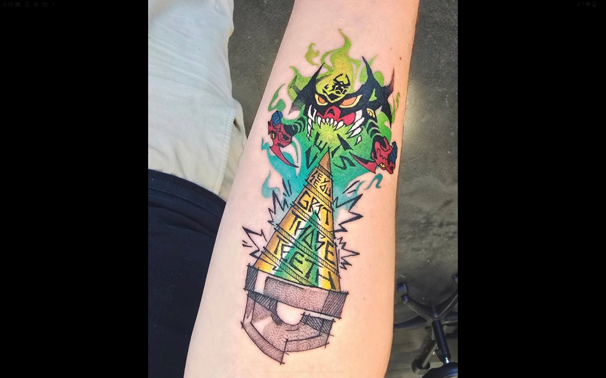 Colorful tattoo on a person's arm depicting a weapon with flames and two fierce cartoonish feline faces with a fiery presence above.