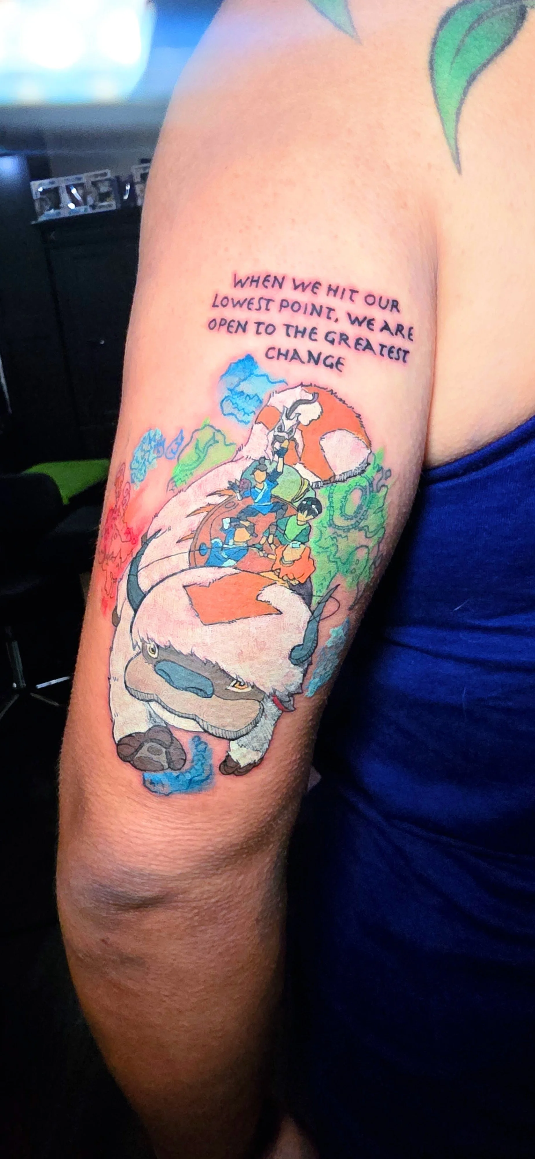 Colorful tattoo on person's upper arm depicting a group of characters riding a large creature with mountains and clouds in the background, accompanied by the quote, 'When we hit our lowest point, we are open to the greatest change.'