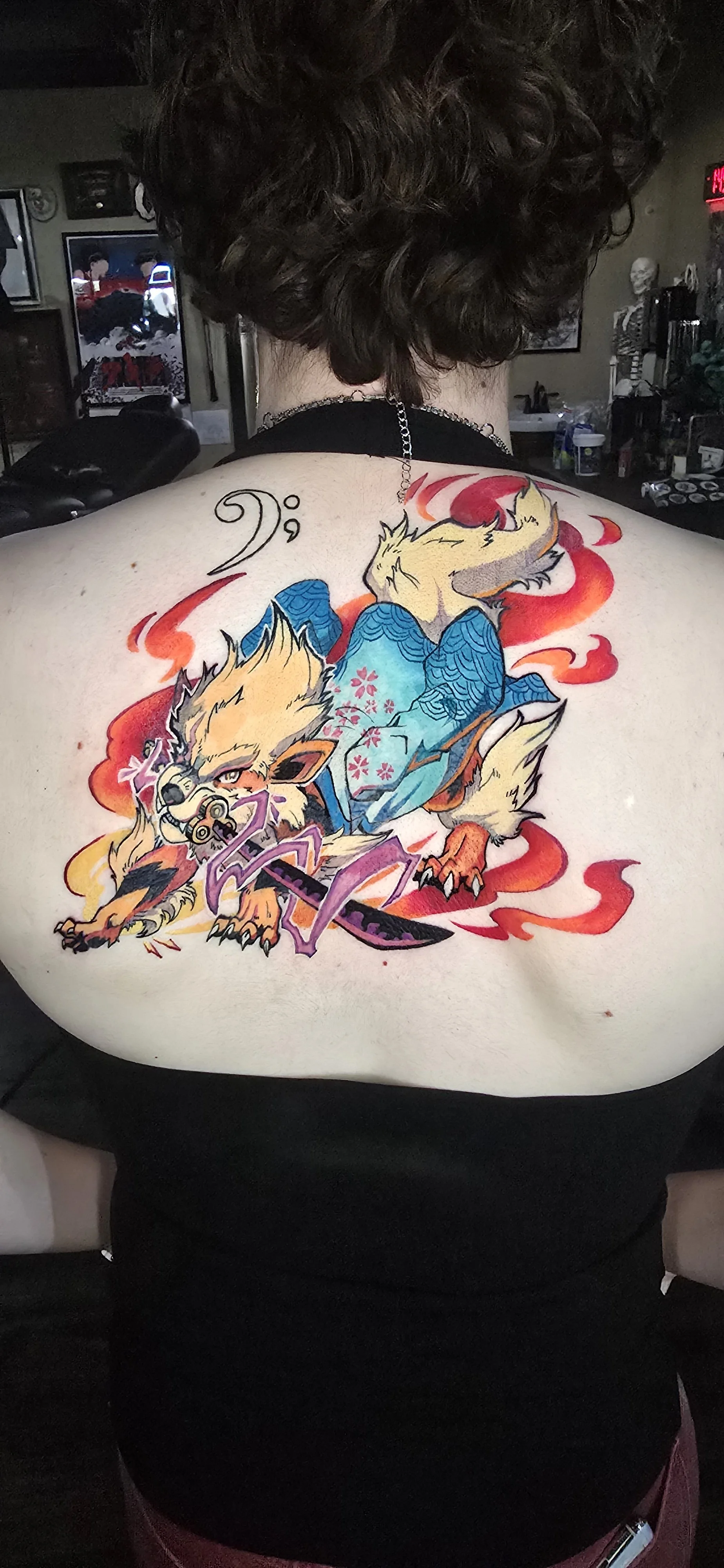 Colorful tattoo of a fox dressed as a samurai, holding a katana, with flames and cherry blossoms around it, on a person's back.