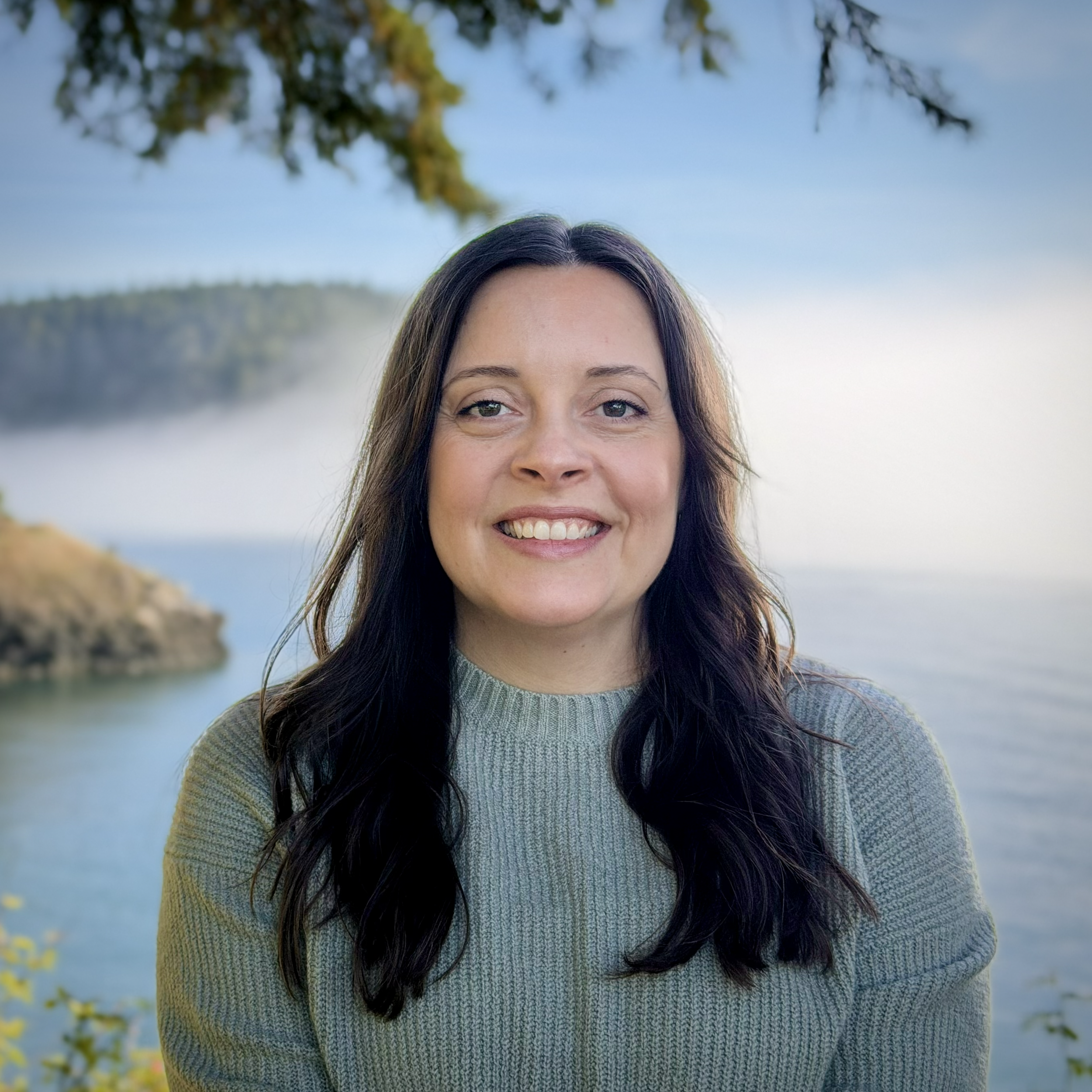 Headshot of Laura Handley smiling outdoors with soft focus water and mountains behind her. On a sunny day with fog over the water.