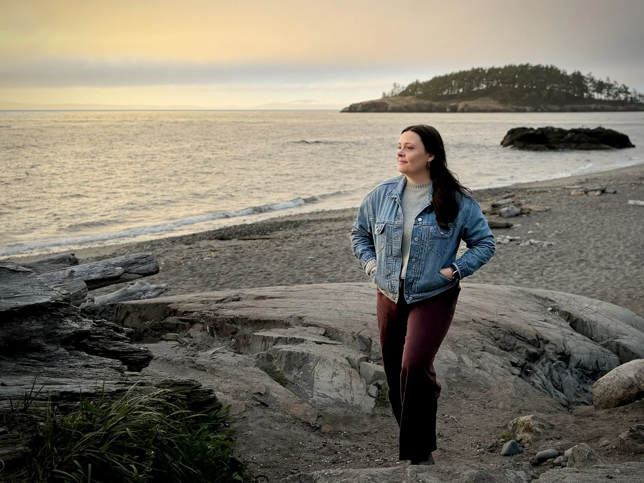 Image of Laura Handley walking along a pacific northwest beach at sunset. Wearing a jean jacket, sage green sweater, and brown pants.