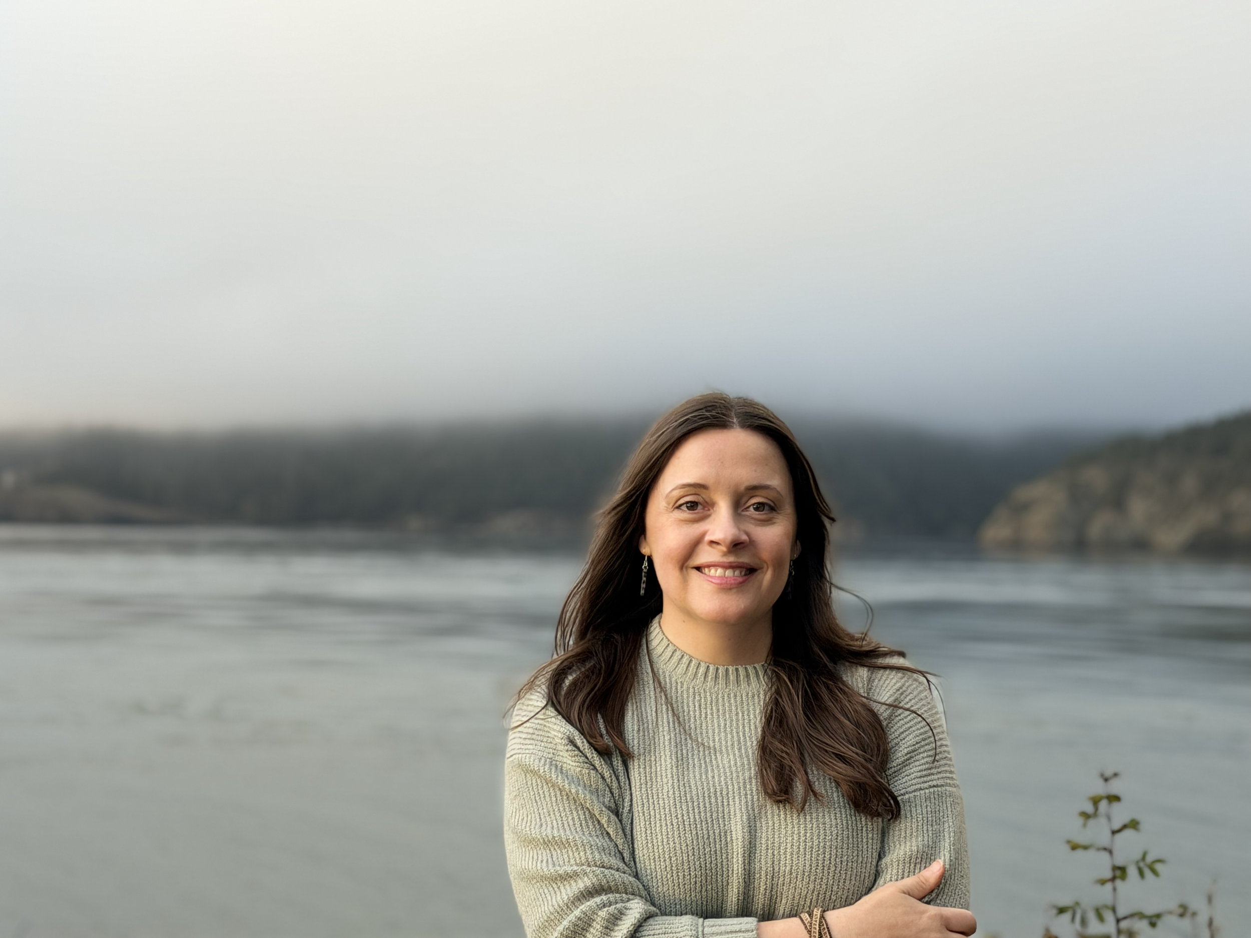Headshot of Laura Handley smiling wearing a green sweater with water and mountains behind her in soft focus on a foggy day.