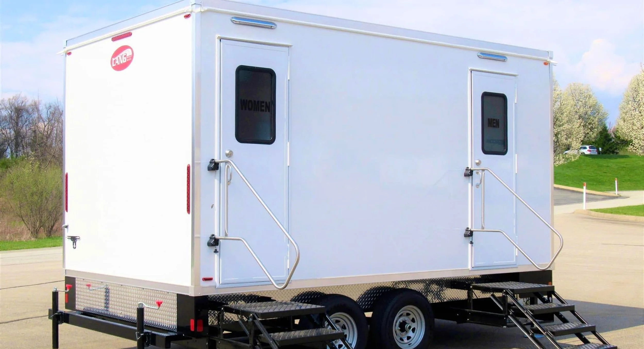 Portable mobile restroom trailer with separate doors labeled 'Women' and 'Men' set outdoors on a paved area with trees and a parking lot in the background.