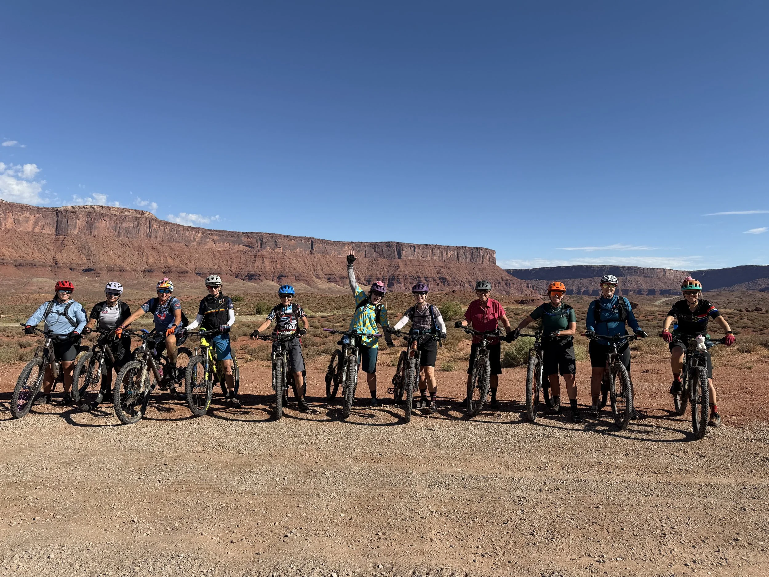 A group of women mountain bikers with mountain bikes standing in a line on a dirt path in a desert landscape with red rock formations under a clear blue sky, riding the Kokopelli Trail.
