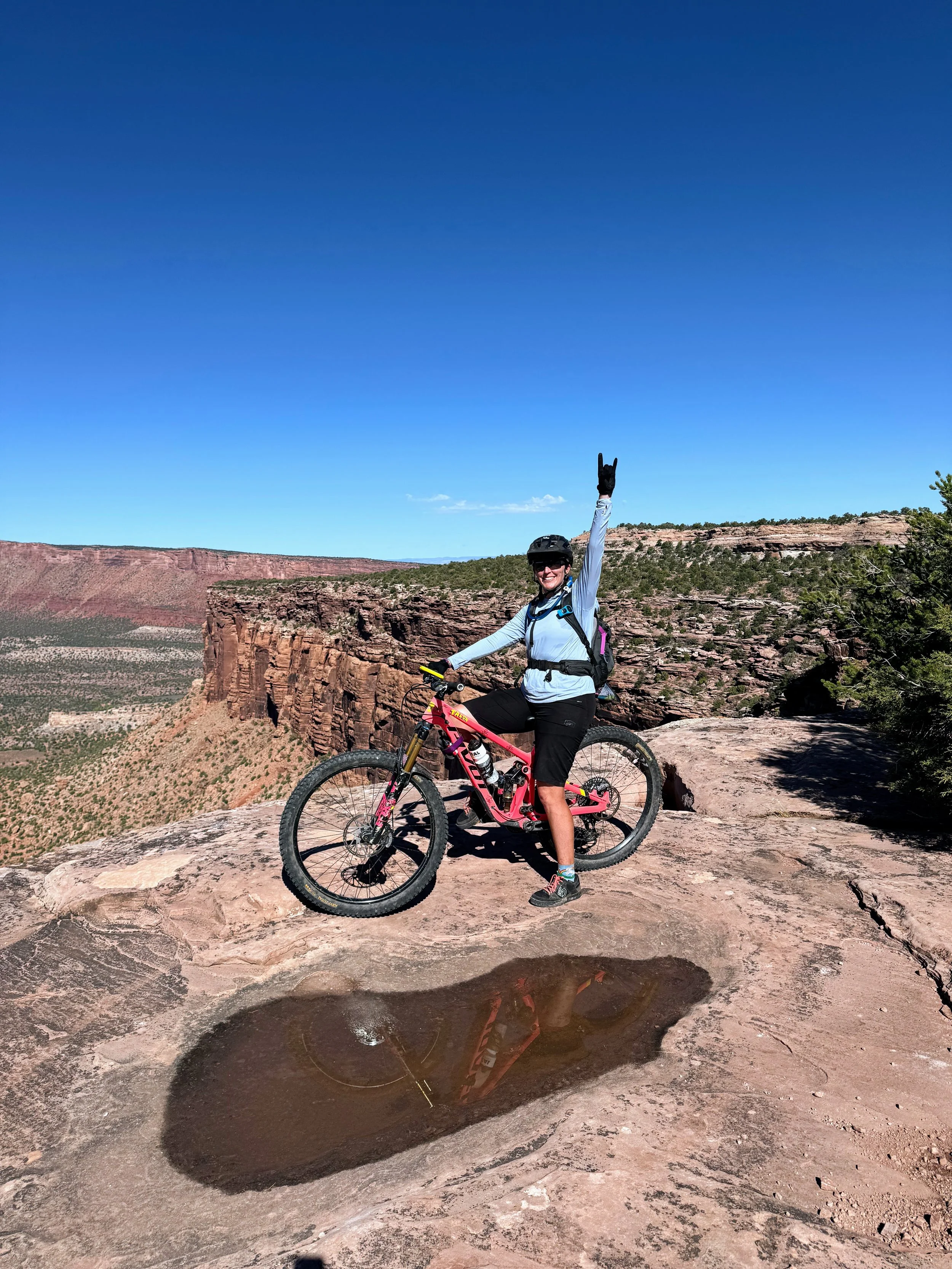 Person in cycling gear standing with a mountain bike on a rock in a canyon landscape with blue sky and red cliffs, a Bikerpelli classic photo