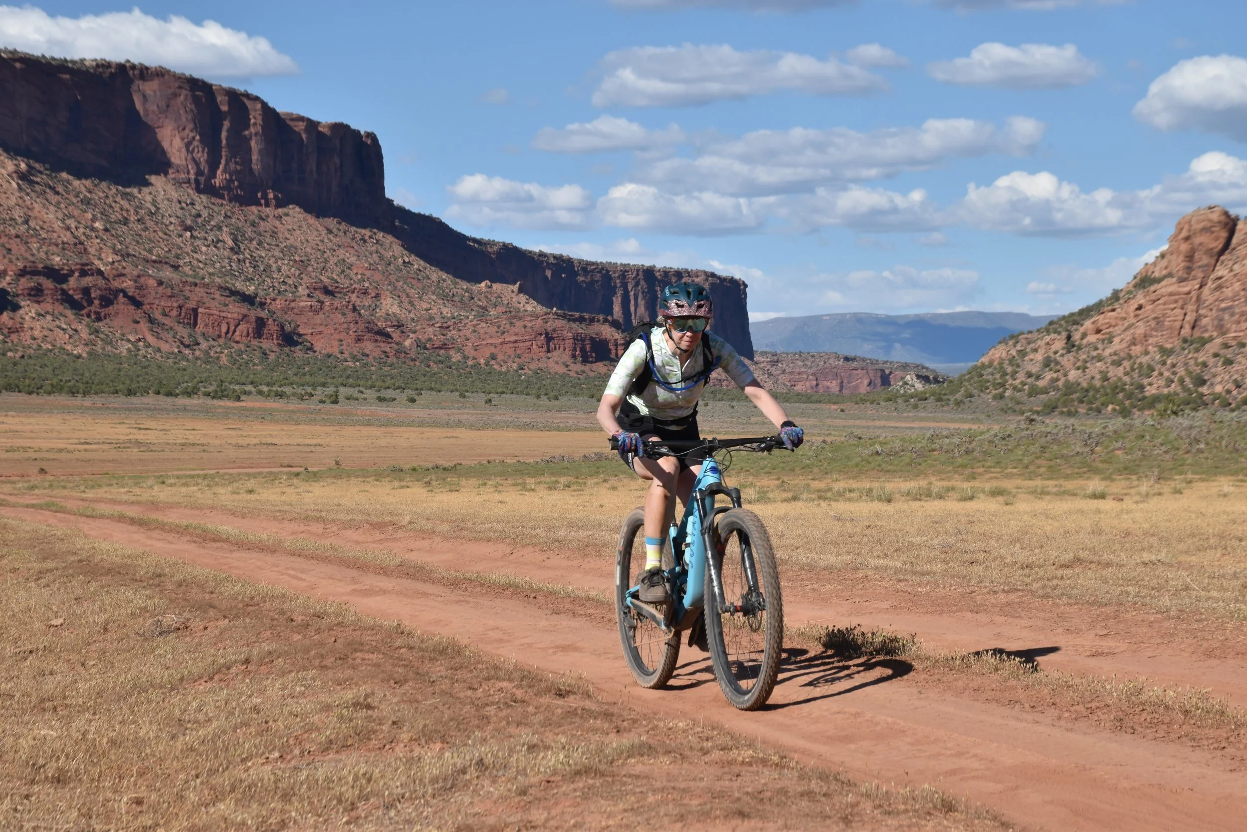 A person riding a mountain bike on a dirt trail through a desert landscape with red rock formations and cliffs under a partly cloudy sky.