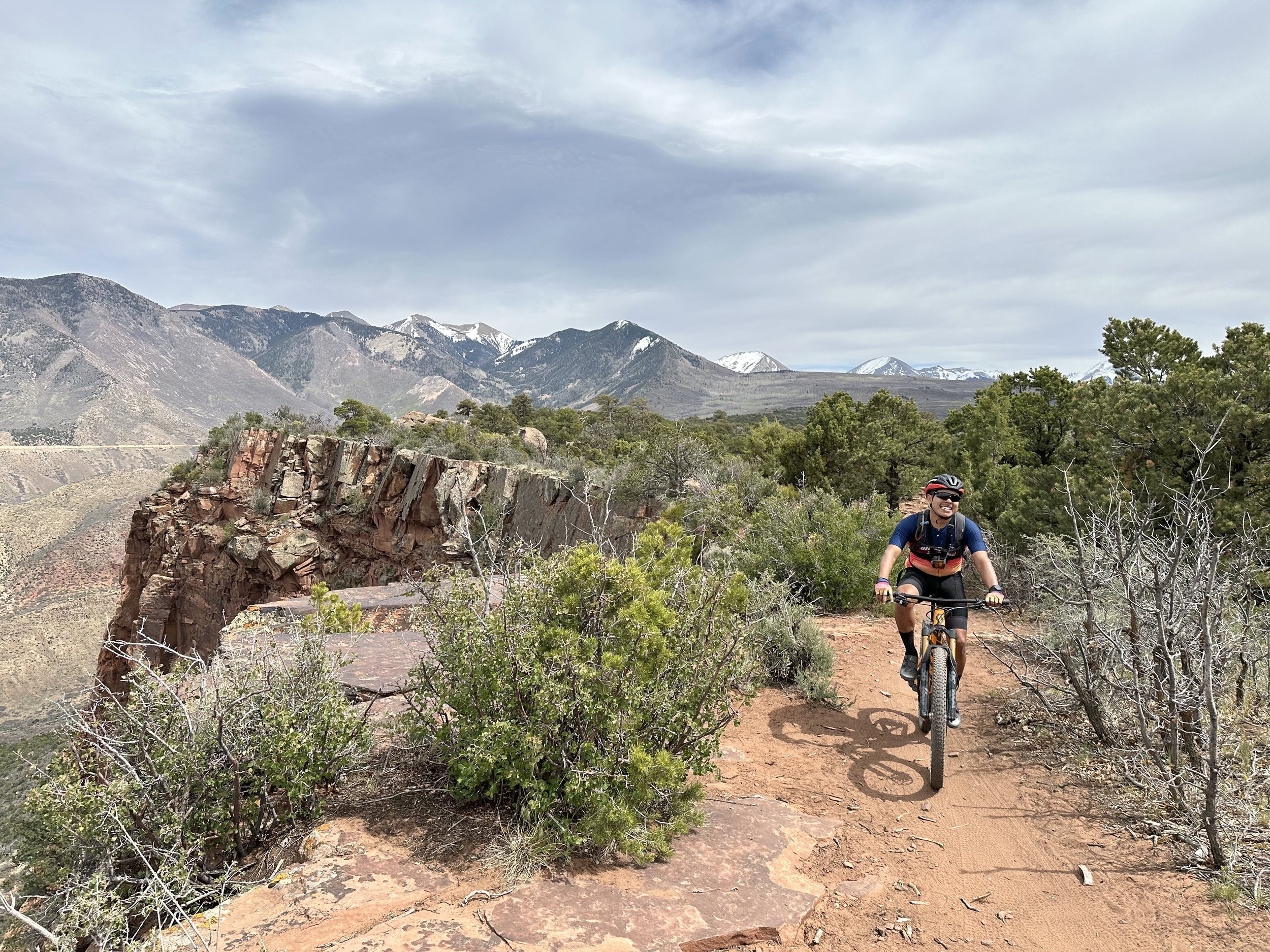 A person riding the Kokopelli mountain bike on a dirt trail in a mountainous area with trees and rocky formations, under a cloudy sky.