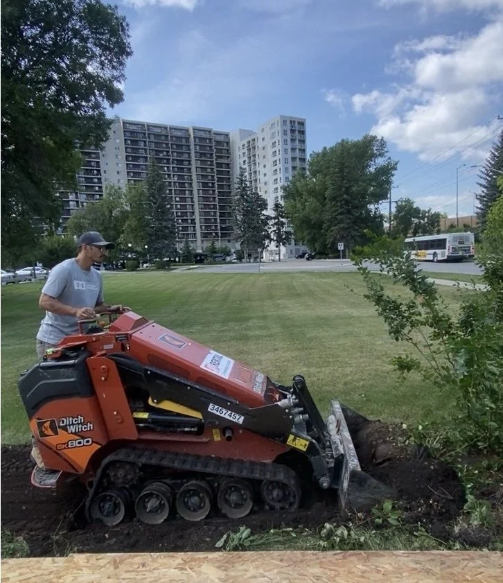 Worker operating a Ditch Witch SK800 compact track loader digging beside a grassy urban park, with apartment buildings and a city bus in the background.