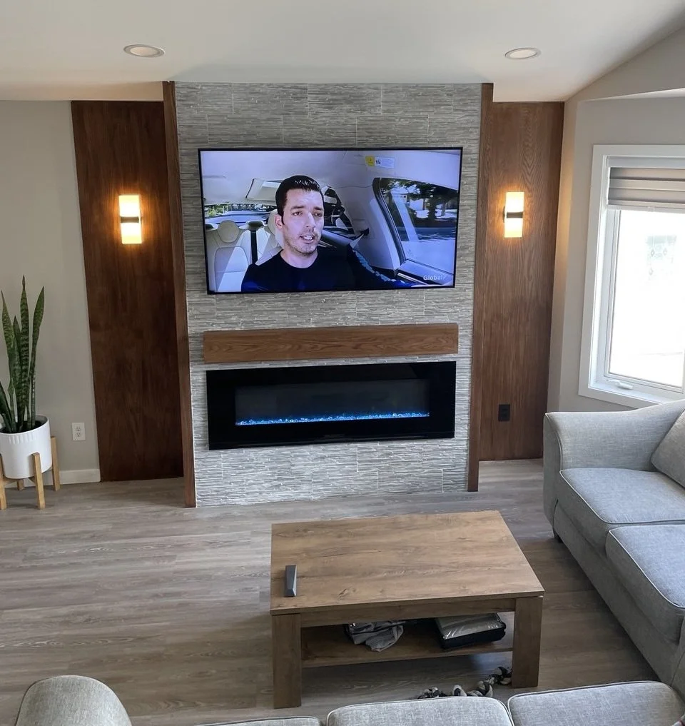 Modern living room with a wall-mounted TV above an electric fireplace, wood paneling, gray stone tile, coffee table, sectional sofa, and potted snake plant