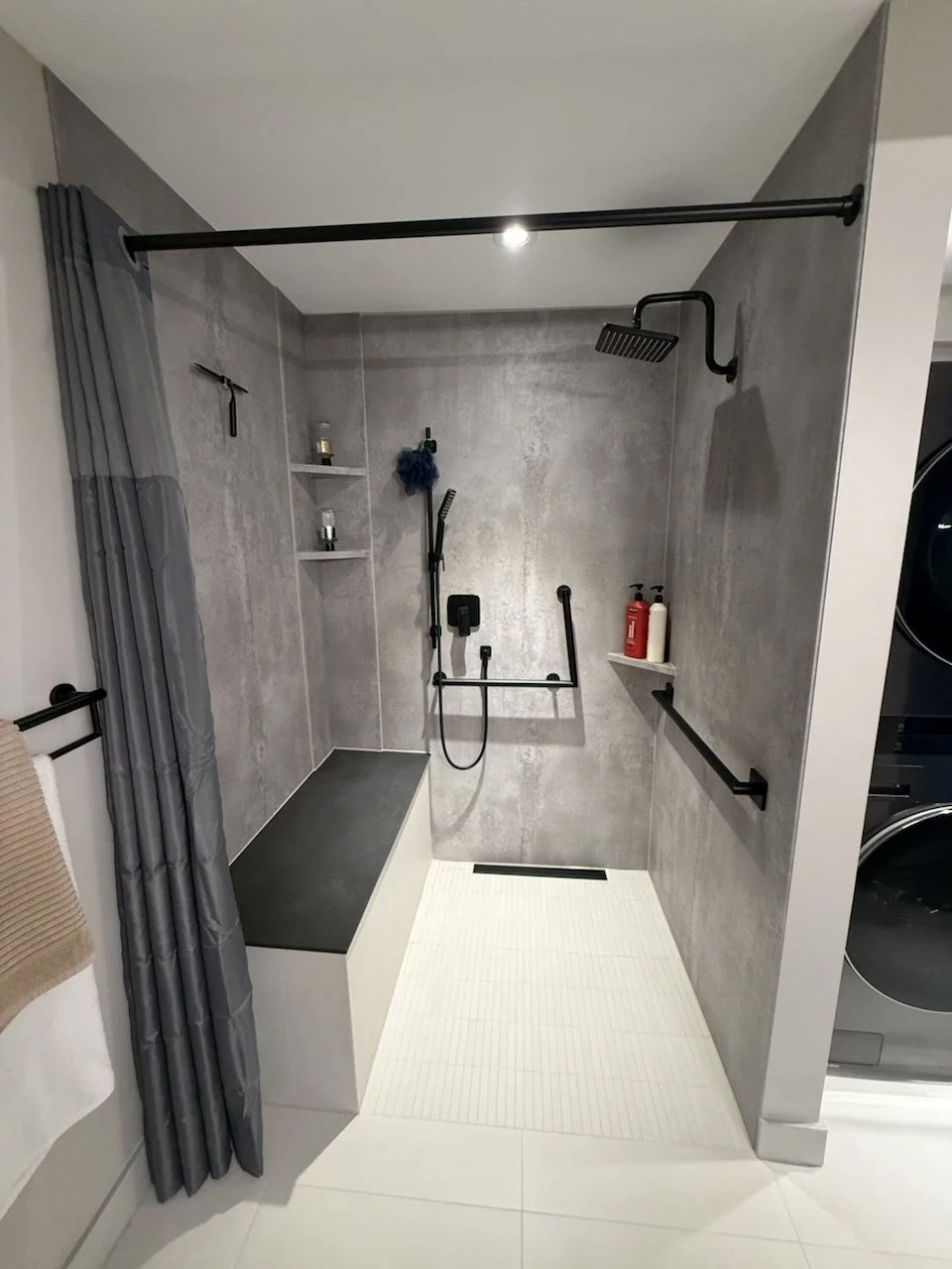 Modern accessible walk-in shower with gray tile walls, black fixtures and grab bars, white textured floor, built-in bench, and gray curtain