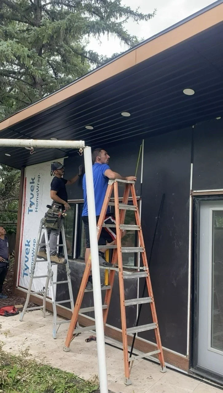 Two contractors installing exterior siding beneath a covered porch roof using step ladders.