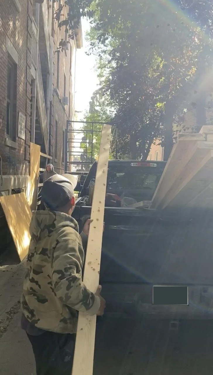 Workers load long wooden boards into a pickup truck beside a brick building.