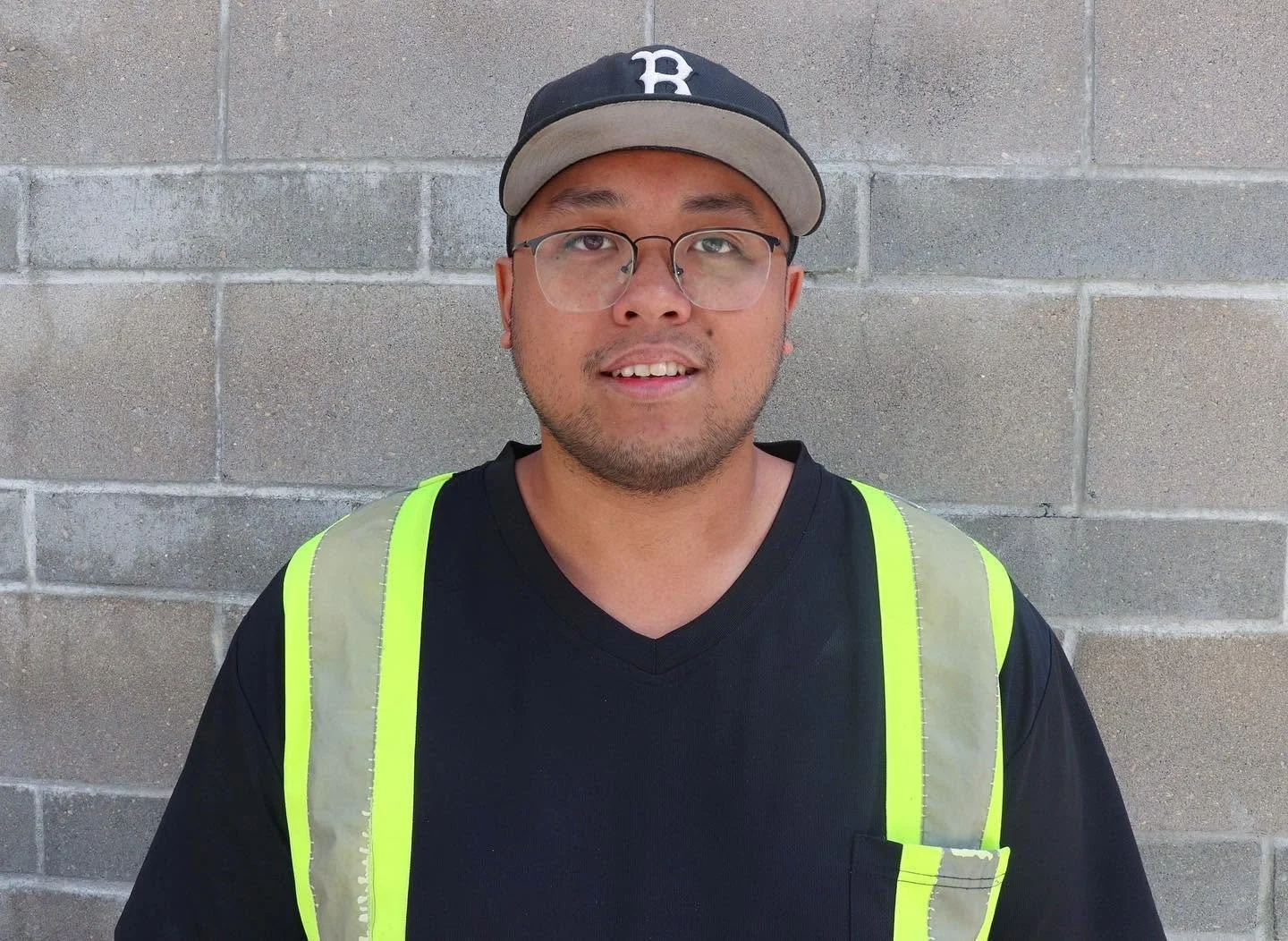 Smiling man wearing glasses, a Boston baseball cap, and a neon yellow safety vest in front of a concrete block wall