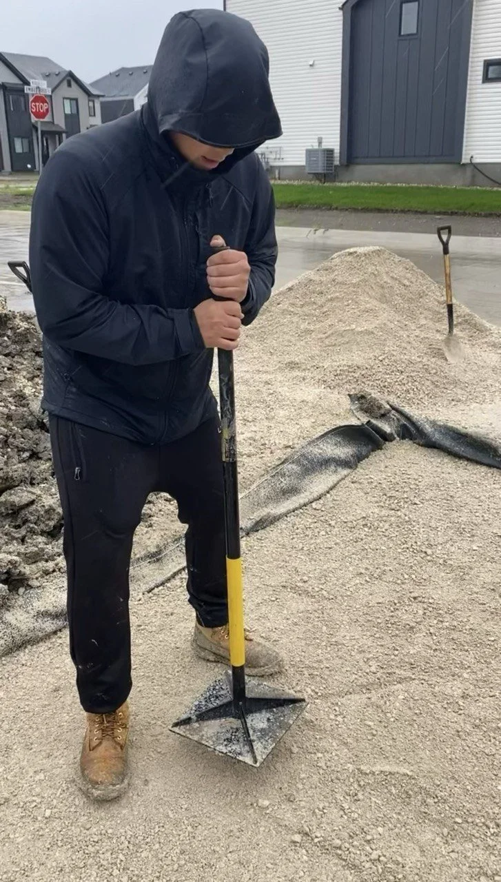 Person in a black hooded jacket spreading gravel with a long-handled landscaping rake on a residential construction site.