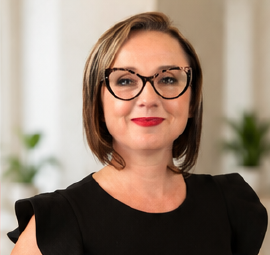 Woman with short brown hair, glasses, and red lipstick in an indoor setting.