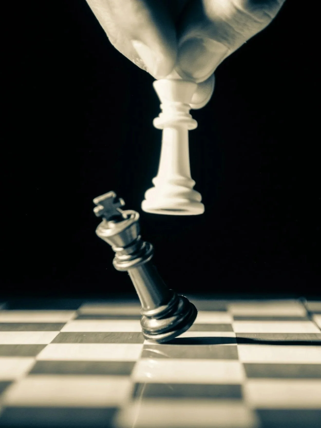 A black and white chessboard with a hand holding a white king chess piece and a black king chess piece falling over.