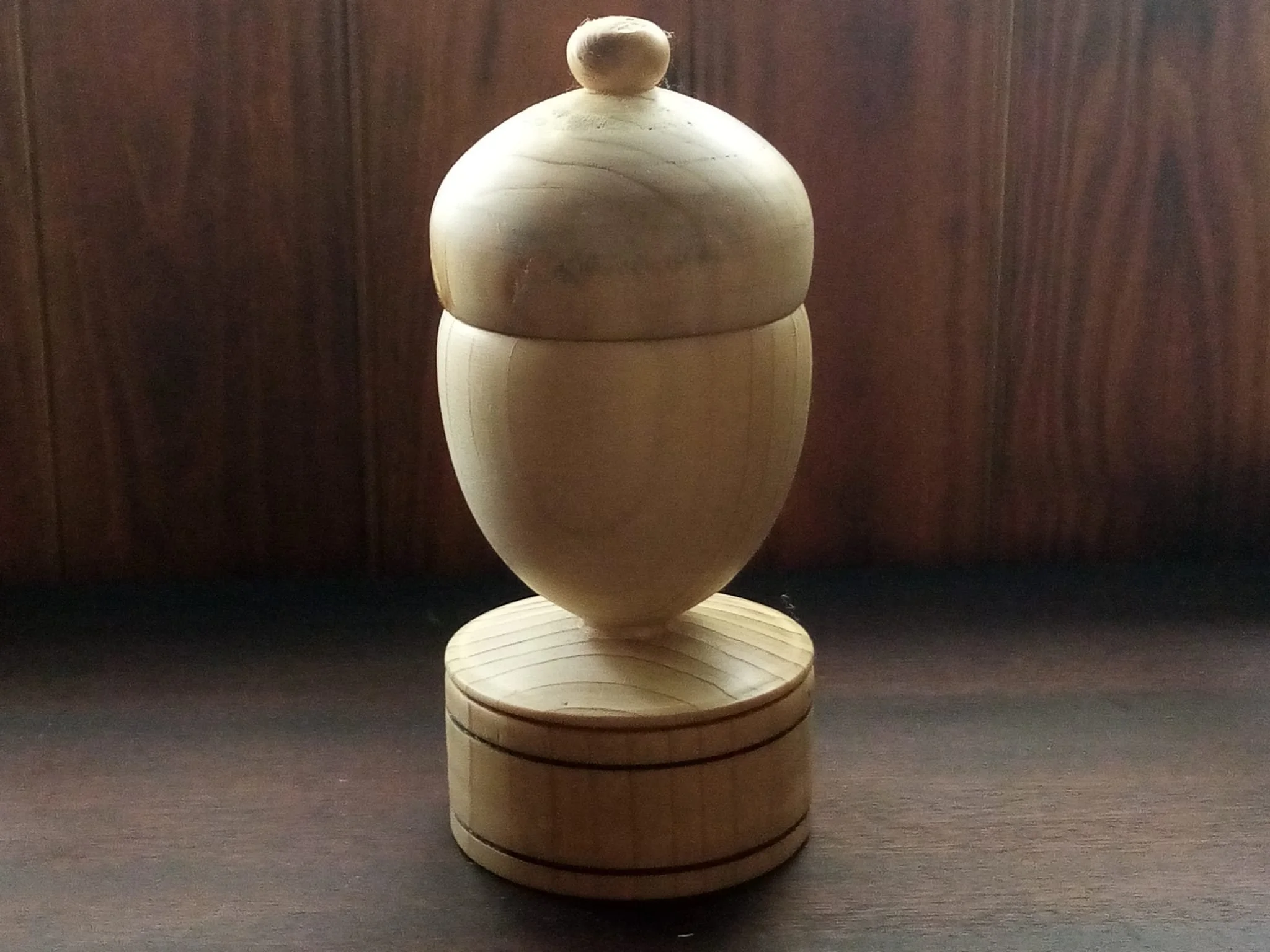Joyero de madera clara en forma de bellota, con tapa rematada en un pequeño pomo y base cilíndrica con dos líneas oscuras cerrado