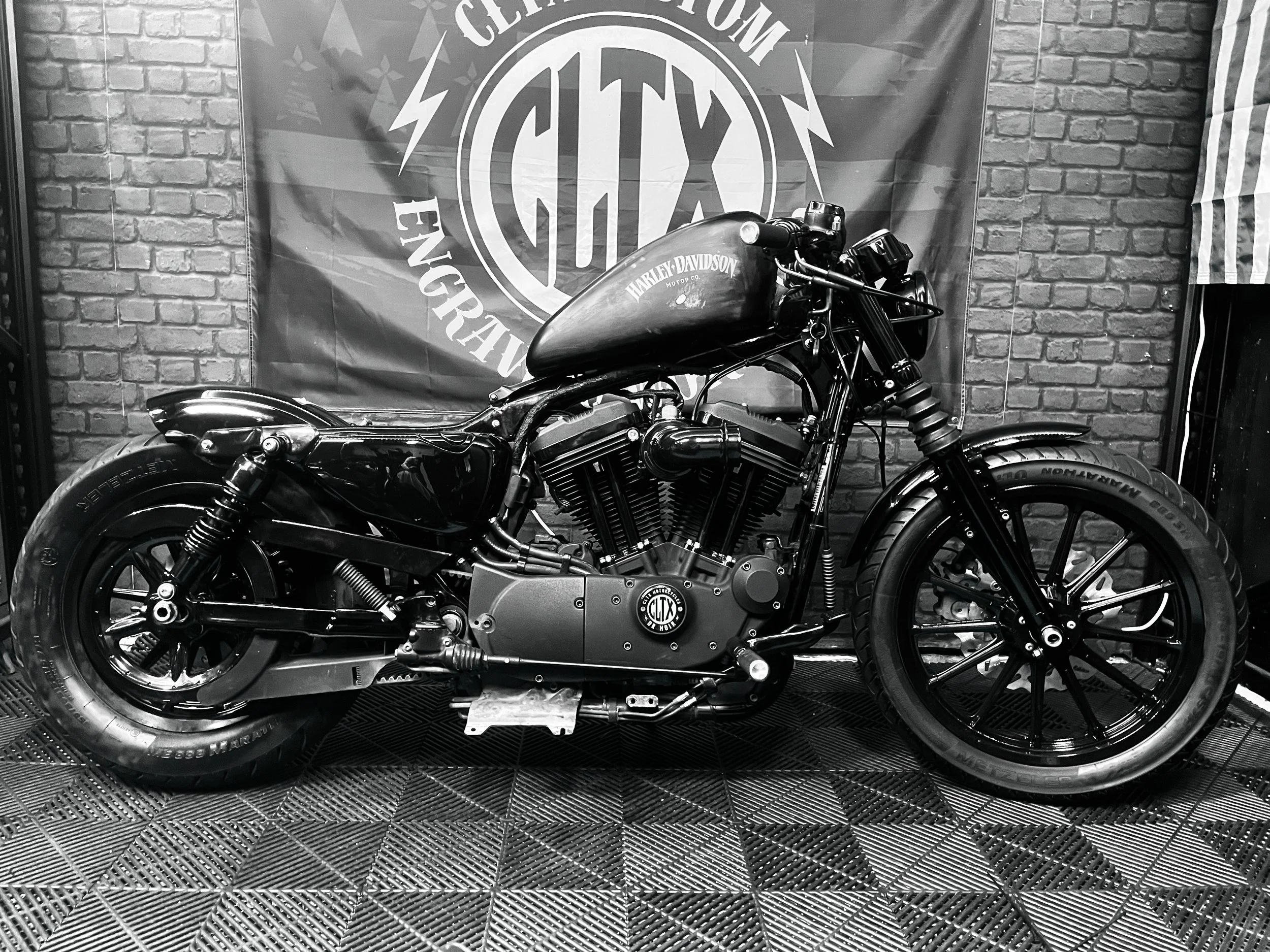 Une moto Harley Davidson noir de style custom est exposée contre un mur en briques, avec une bannière du club de construction de motos sur le fond.