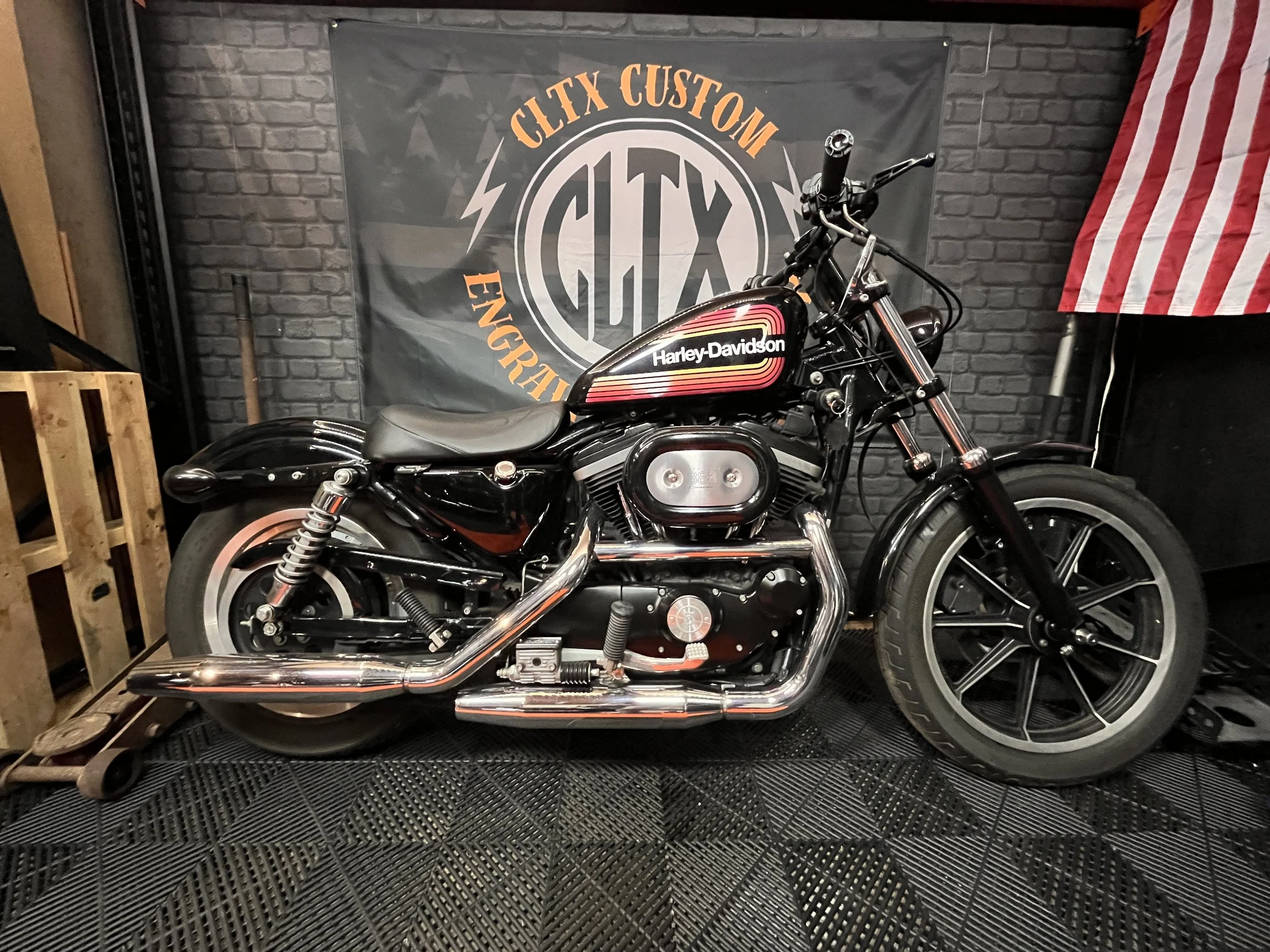 Une moto Harley-Davidson noire est affichée devant un mur en briques avec une bannière Grltx Custom.