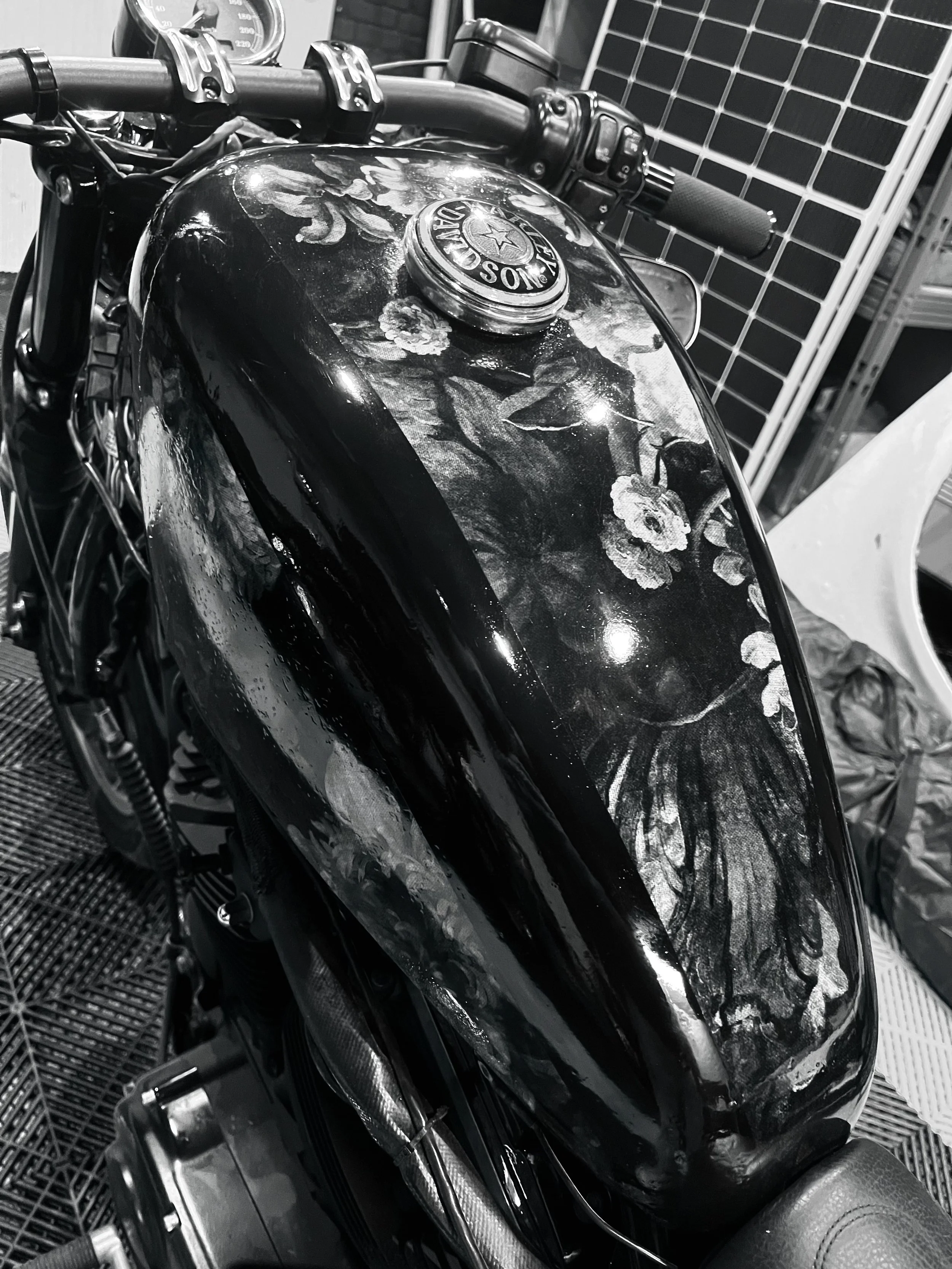 Une moto avec un réservoir décoré de fleurs, vue en gros plan, en noir et blanc, avec les détails du guidon, du tableau de bord et des câbles visibles.