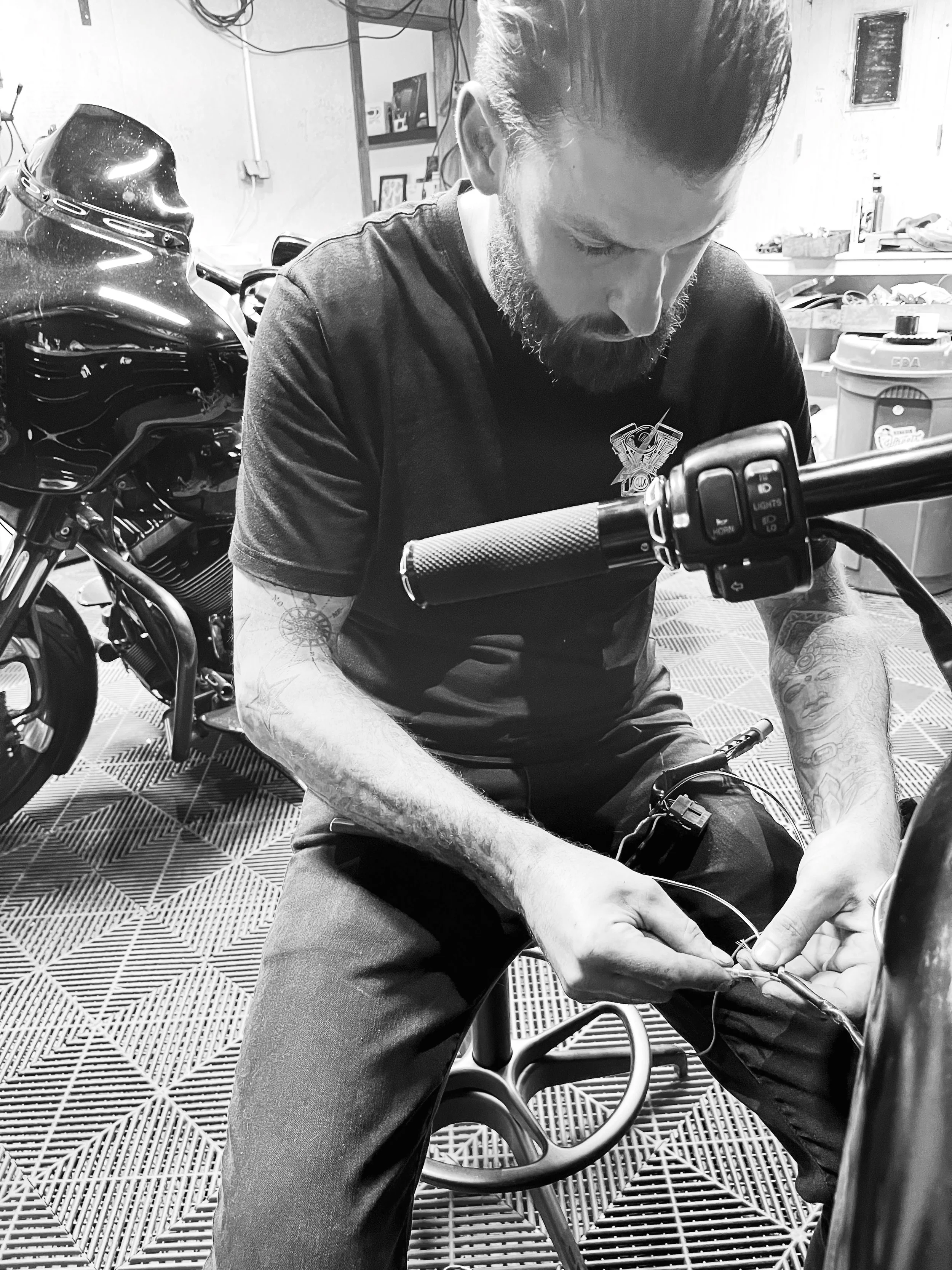 Un homme avec des tatouages, portant un t-shirt noir, œuvre sur la moto dans un garage.