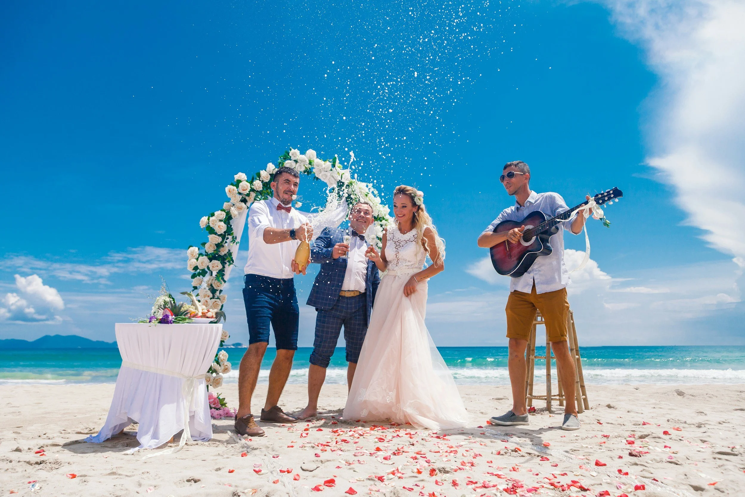 Beach wedding celebration with a bride, groom, friends, champagne spray, floral arch, and guitarist by the ocean