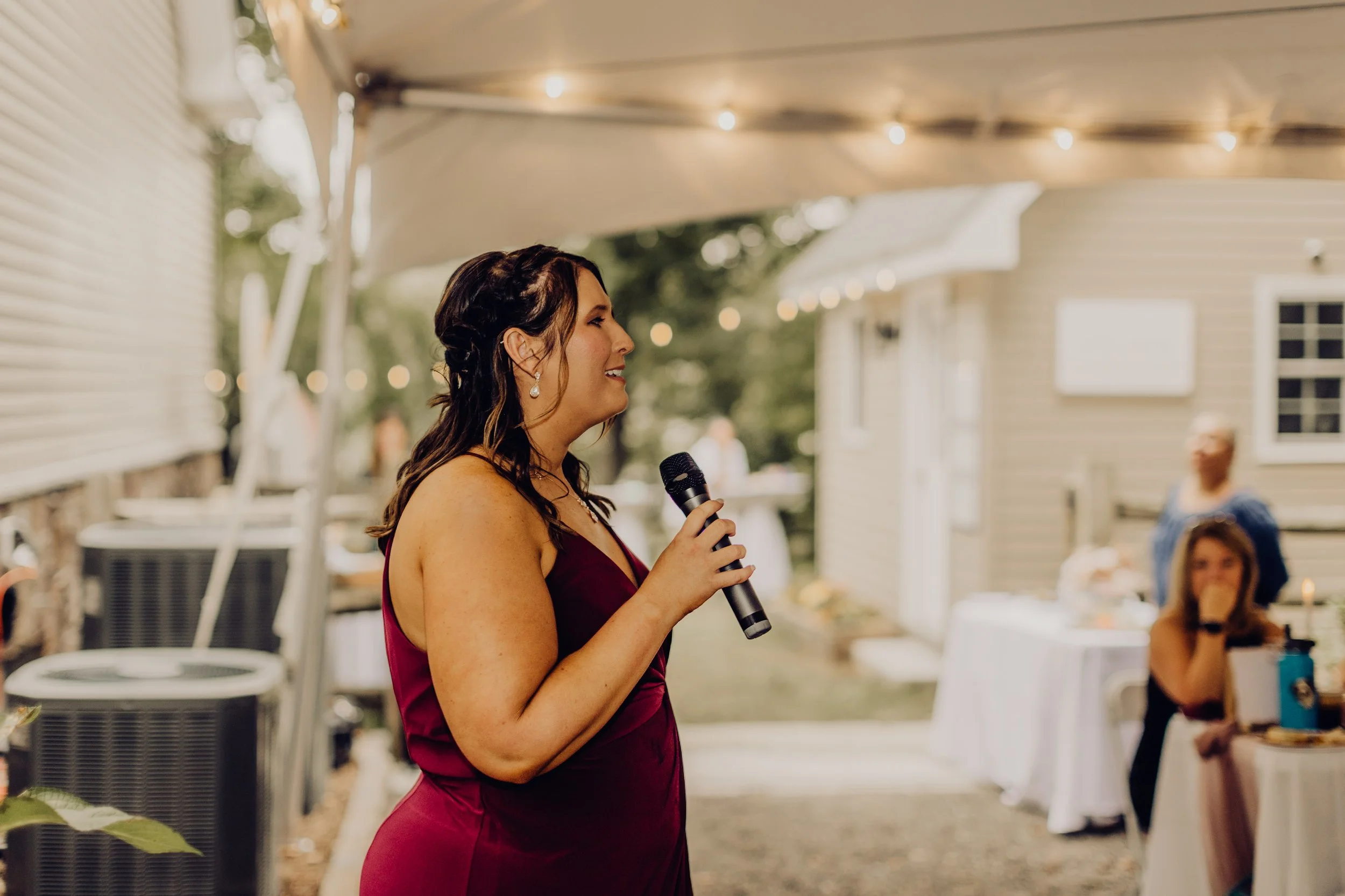 Woman in a burgundy dress singing into a microphone at an outdoor party with string lights and guests.