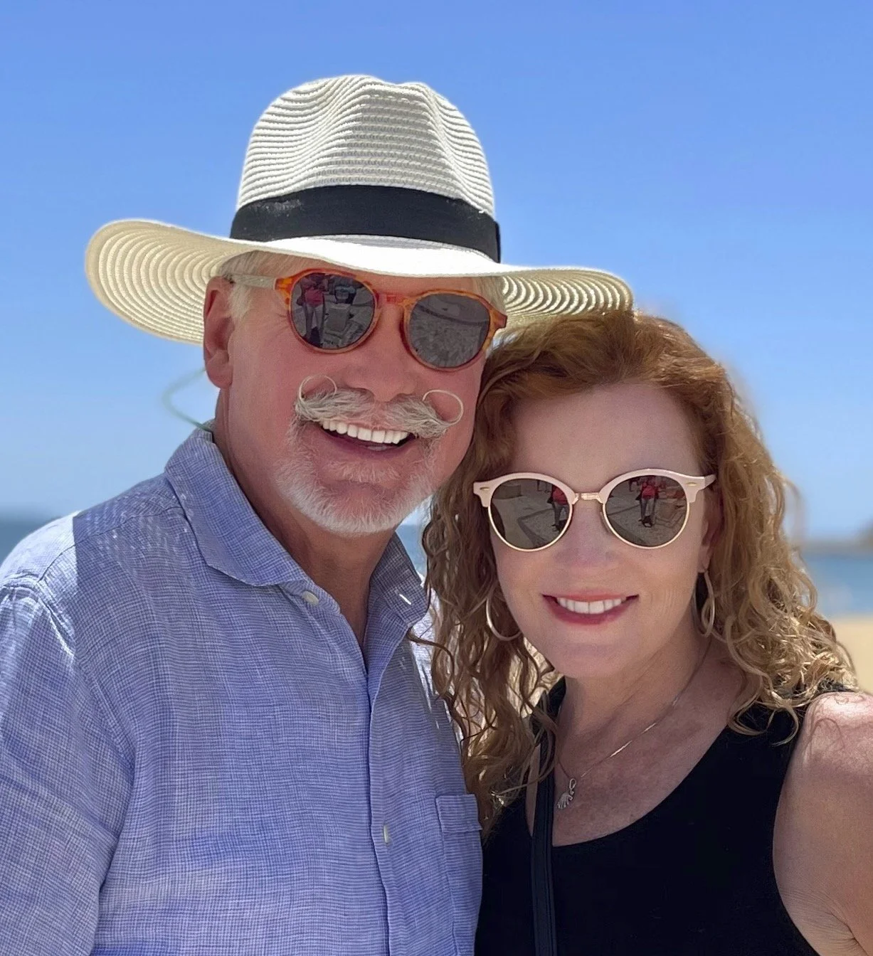 Photographer Greg Cavanagh and his wife Cheryl smiling together outdoors beneath a clear blue sky.