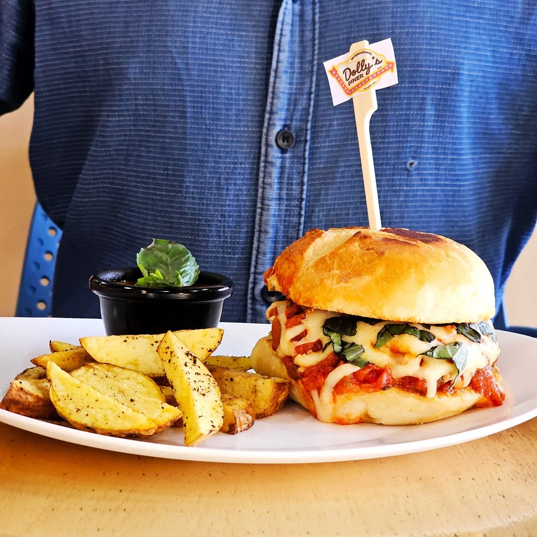 Hamburguesa con queso, tomate y espinaca, acompañada de papas fritas y salsa en pequeño bol, en un plato blanco en una mesa, con una persona de camisa azul en el fondo.