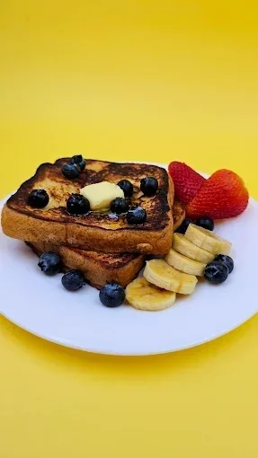 Dos rebanadas de pan tostado con mantequilla, cubiertas con arándanos y acompañadas de plátano en rodajas y fresas, en un plato blanco sobre un fondo amarillo.
