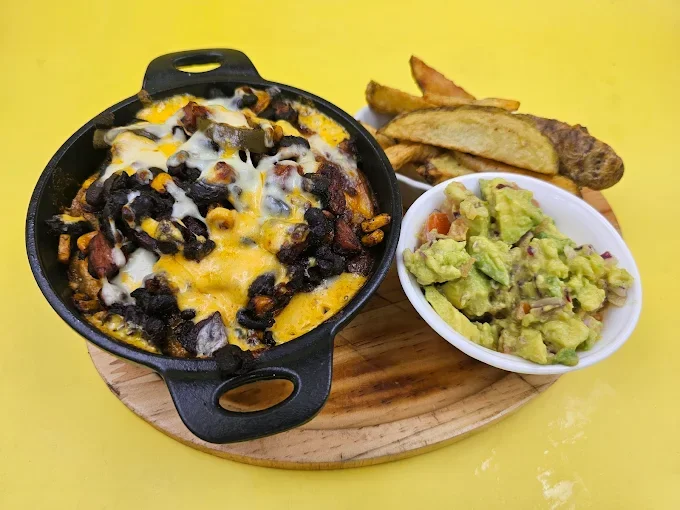 Porción de nachos con queso, frijoles negros y carne, acompañados de guacamole y papas fritas, sobre una tabla de madera en un fondo amarillo.
