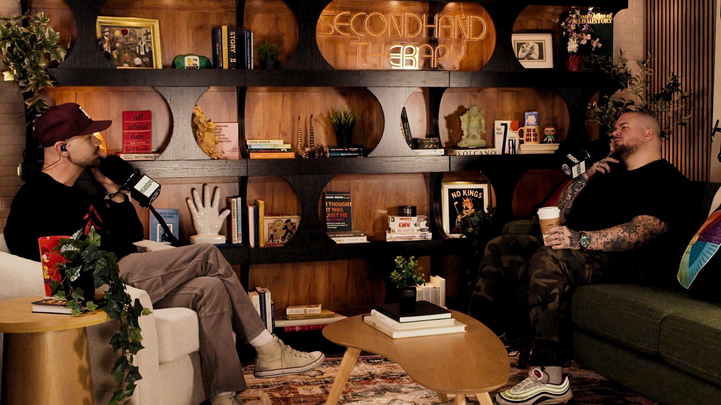 Two men seated indoors, talking into microphones during a podcast or interview, with a wooden bookshelf behind them filled with books, plants, and decorative items. Sign on shelf reads 'Second Hand Therapy'.