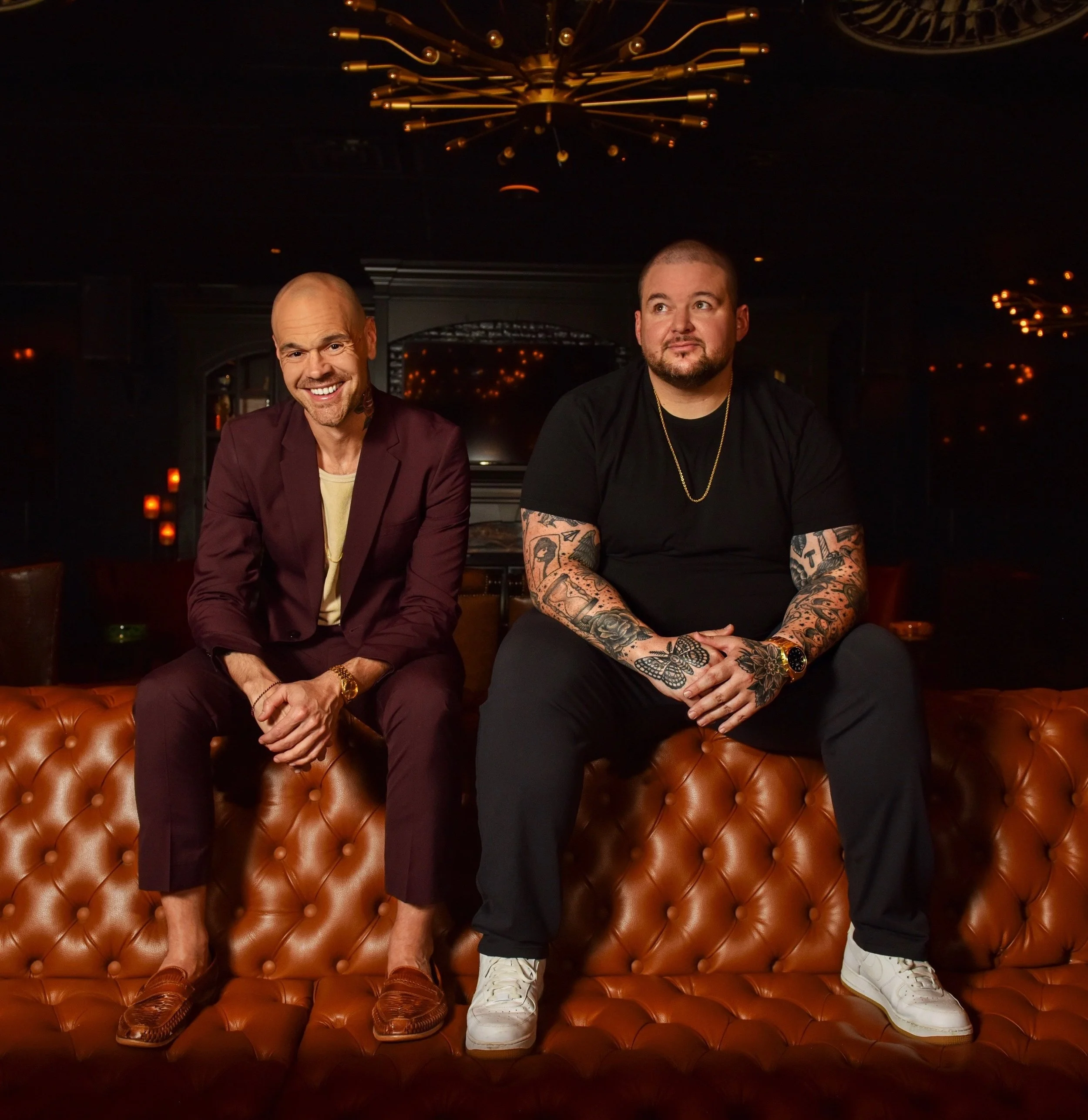 Two men sitting on a tufted leather bench in a dimly lit lounge or bar with modern chandeliers and warm lighting.
