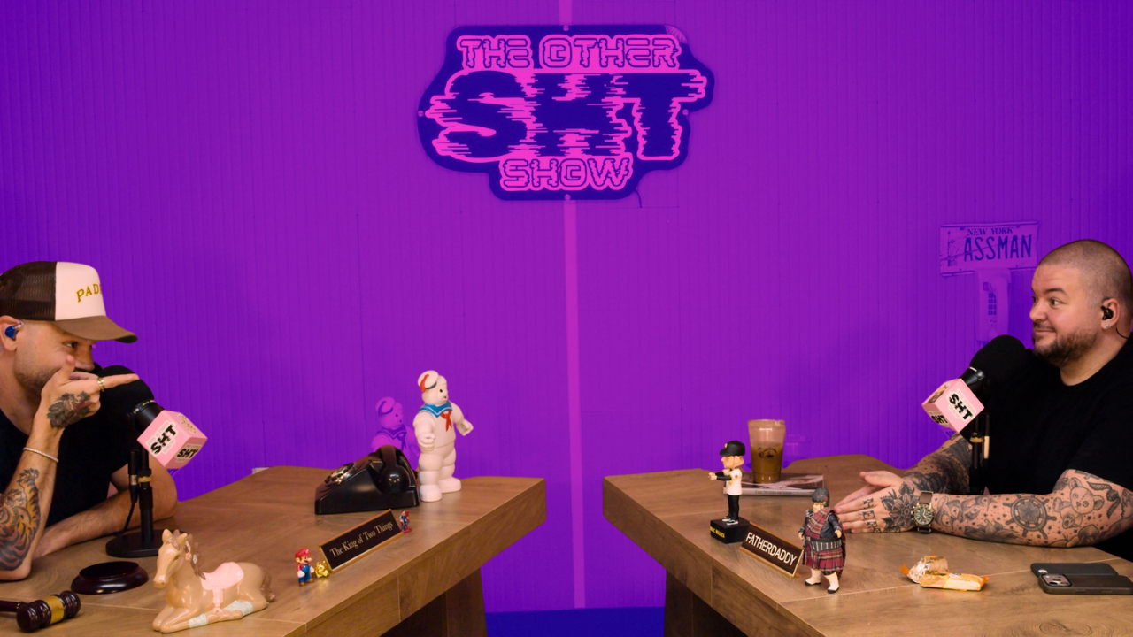 Two men are sitting at a wooden table in a podcast studio with purple walls. There is a sign above them that reads "The Other Sht Show" in neon. The man on the left is wearing a cap that says "PADD" and has tattoos on his arm. The man on the right has tattoos on his arms and is smiling. The table has various collectibles including a plush Soviet-era figure, a small Mario figurine, and figurines labeled "The King of the Things" and "FatherDADDY." There is a glass of iced coffee in front of the man on the right.