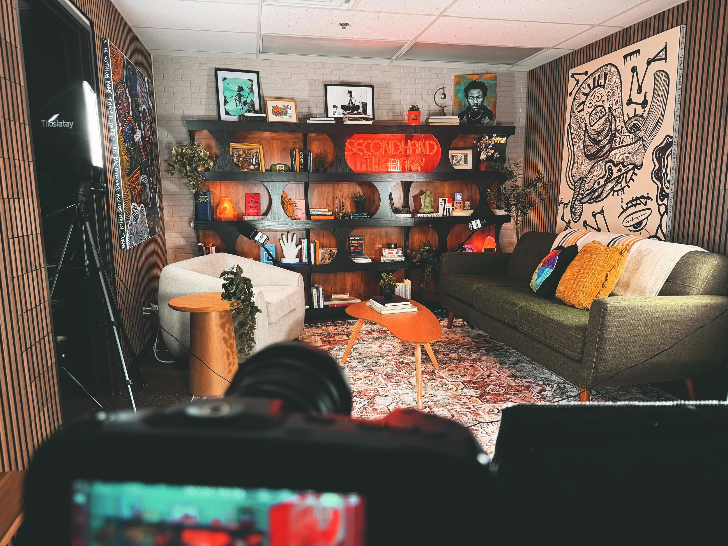 Interior of a cozy living room with a large wooden bookshelf decorated with artwork, plants, and books. There are two chairs and part of a sofa with colorful pillows, a patterned rug, and artwork on the walls. A neon sign on the bookshelf reads 'SECONDHAND THERAPY.'