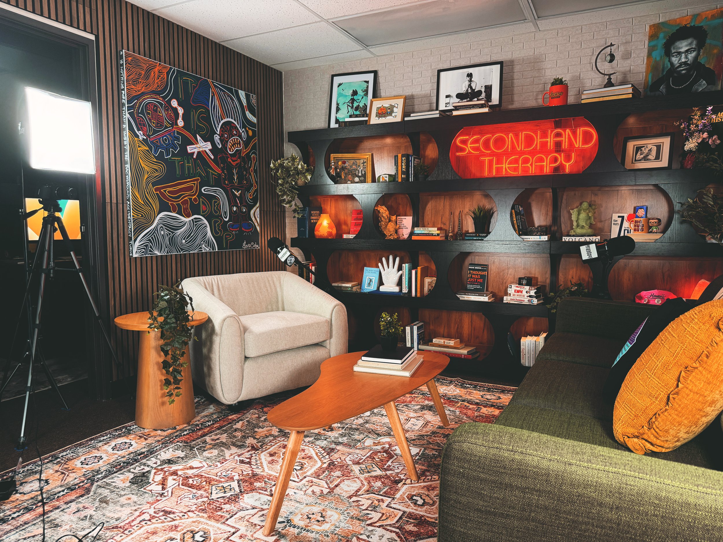 Cozy reading room with a white armchair, wooden coffee table, bookshelves with art and plants, a colorful painting, and neon sign reading 'SECONDHAND THERAPY' on the wall.