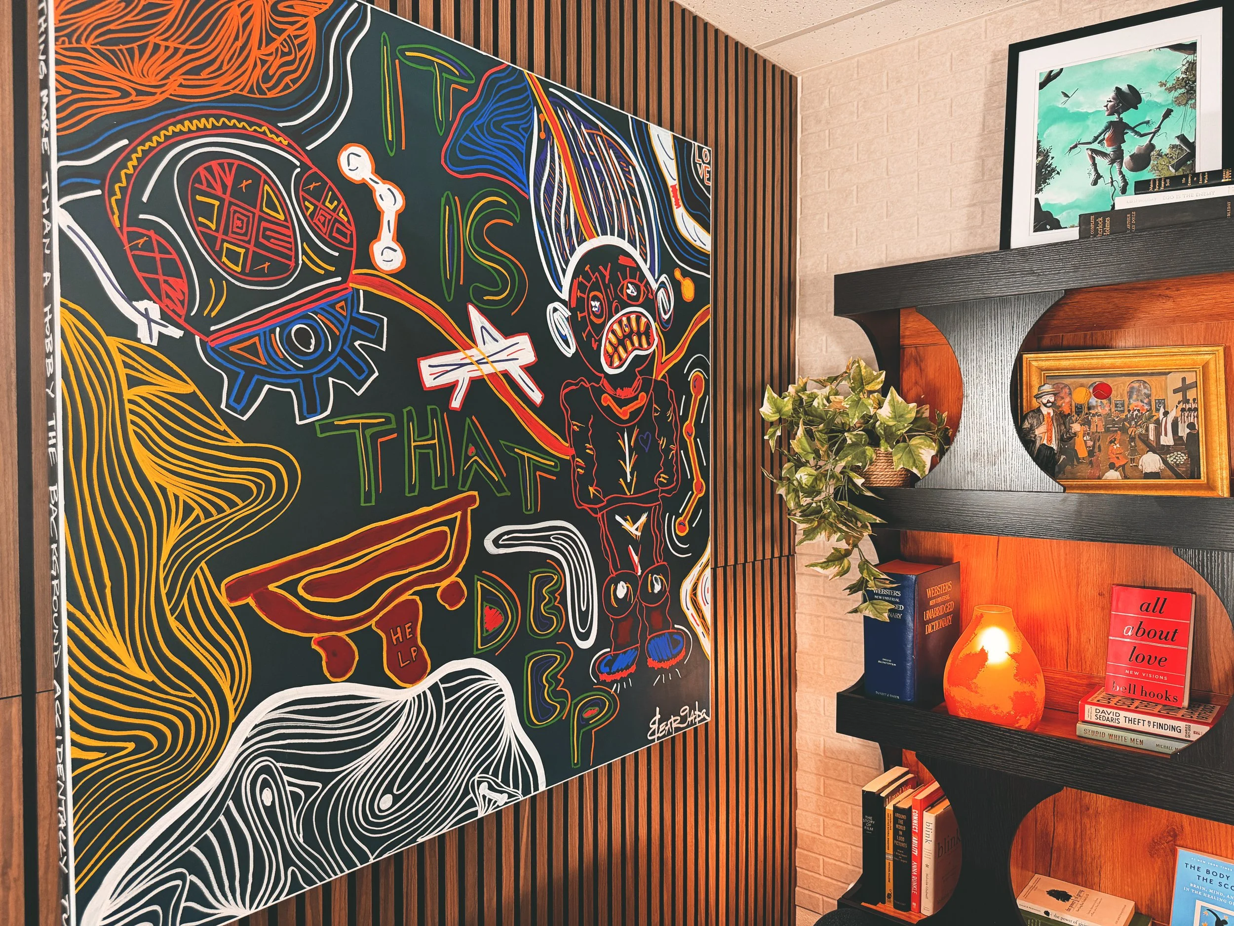 Colorful abstract artwork with various shapes and patterns hanging on a wooden-paneled wall in a cozy room, with a black shelf displaying books, a framed picture of a character, and a decorative orange vase with green plants.