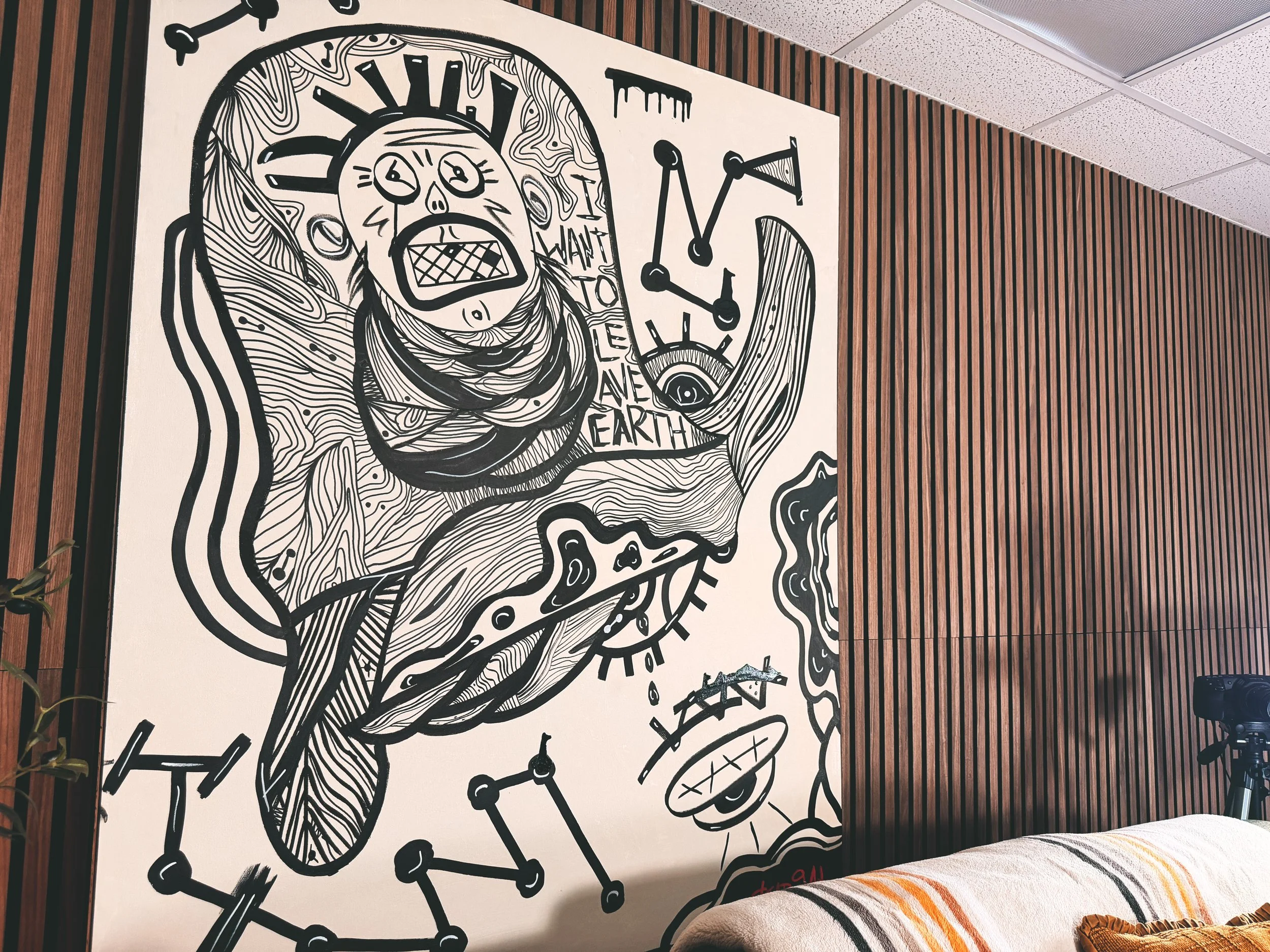 A black and white line art mural features a stylized skull with a crown, surrounded by various abstract symbols, arrows, and patterns, on a wall with wooden paneling.