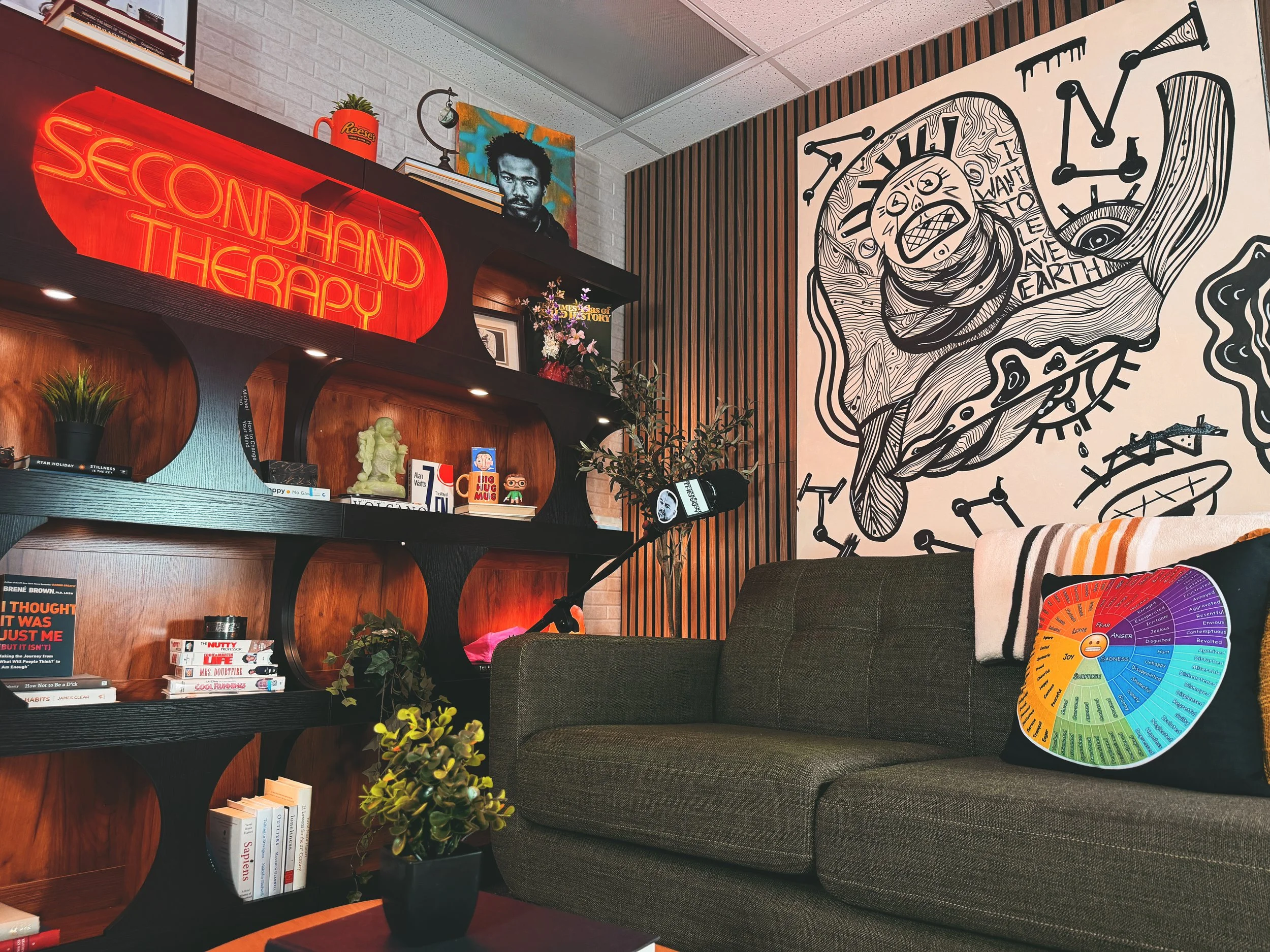 A cozy living room corner with a black bookshelf filled with books, plants, and decorative items, a black and white mural art of a human head with gears and clocks, a dark gray sofa with a colorful wheel chart pillow, a colorful painting of a person's face, and a neon sign reading 'SECOND HAND THERAPY' in red.
