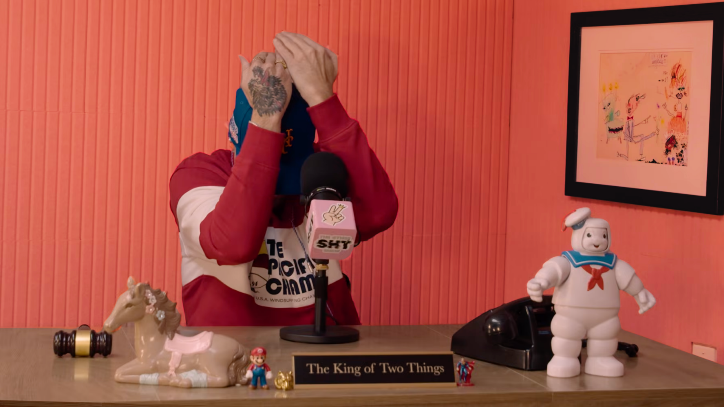 A man wearing a red and white jacket stands at a table with various toys, including a unicorn figure, a Mario figure, a Ghostbusters Stay Puft marshmallow man, and a wooden gavel. The man covers his face with his hands, and there is a framed colorful drawing on the orange wall behind him. A microphone with a pink cover labeled 'SHIT' is in front of him, and a sign reads 'The King of Two Things'.