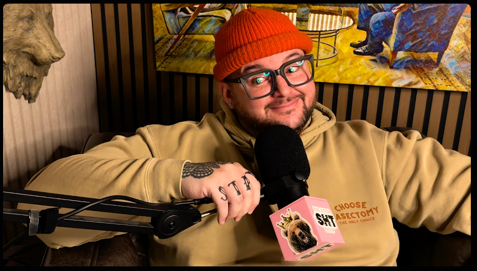 A man with glasses, a red beanie, and a beige hoodie sitting on a couch, holding a microphone with a pink cover featuring a bear. There is artwork of people in a room hanging on the wall behind him.