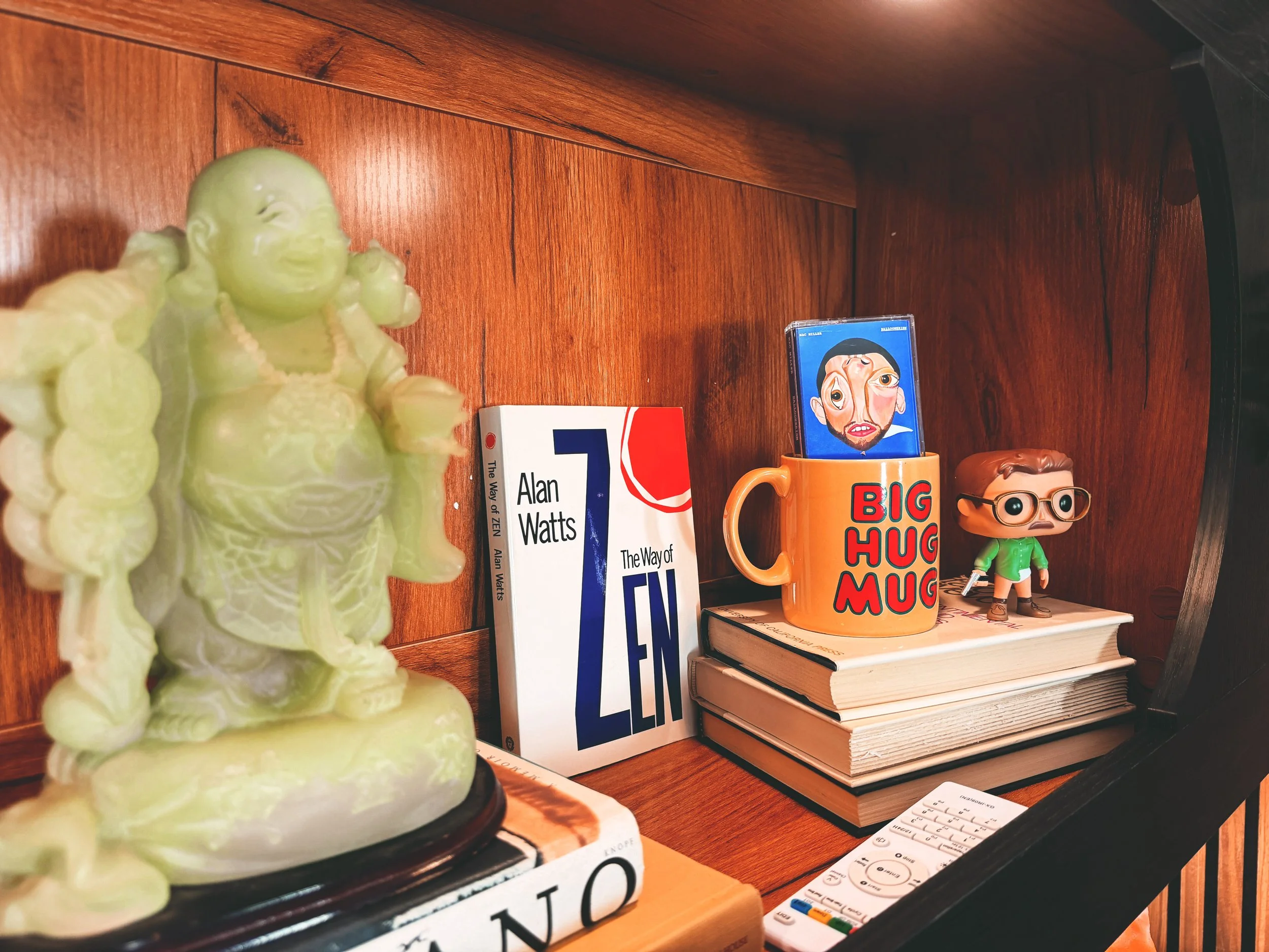 Decorative green Buddha statue on a wooden shelf next to books, a mug with cartoon face, and a small figurine with glasses.