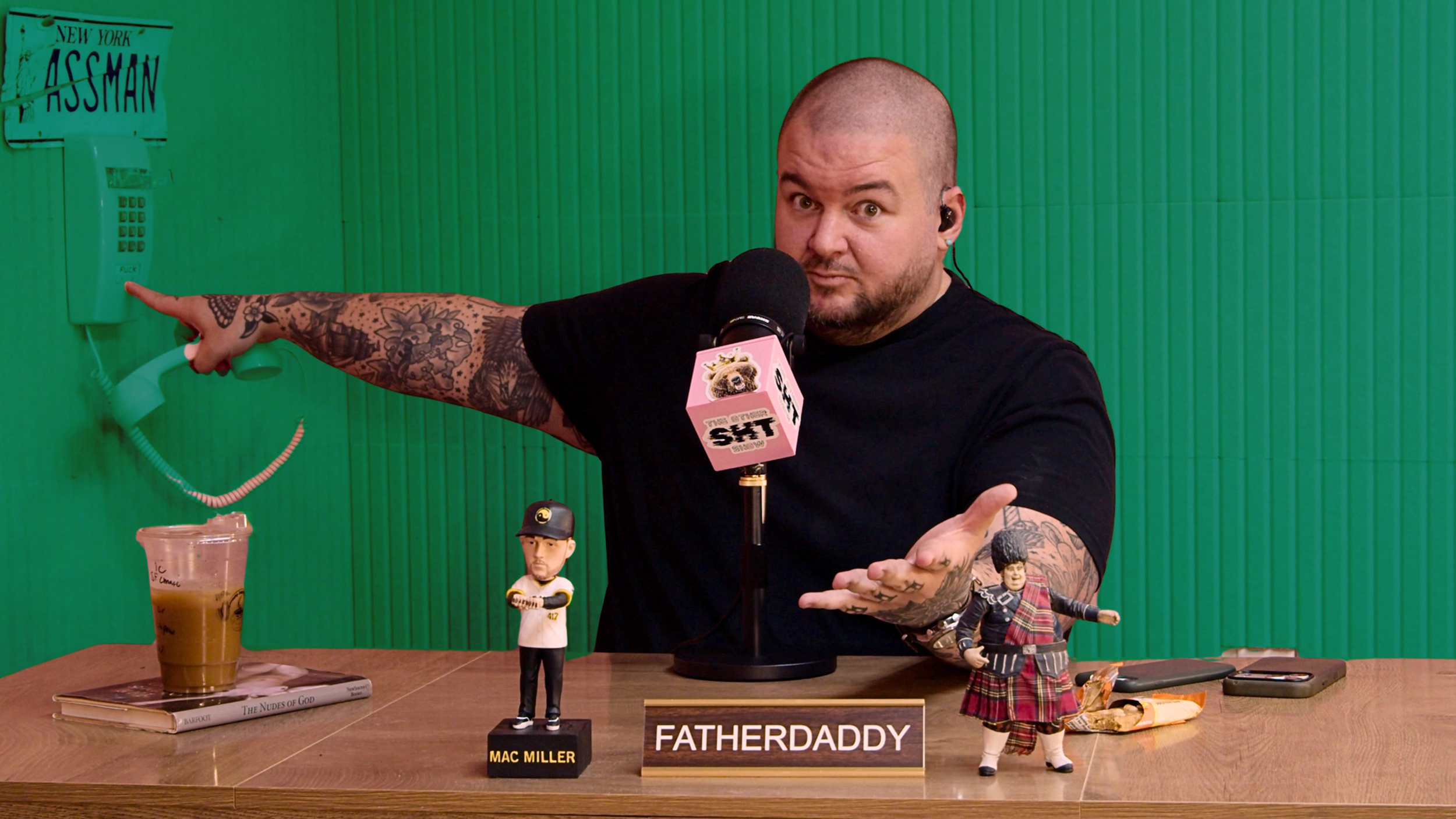 A man with tattoos and a shaved head sits behind a table, pointing with his right hand toward a vintage green payphone mounted on a green wall. In front of him are various figurines and props, including a sign reading 'FATHERDADDY,' a bobblehead labeled 'MAC MILLER,' and figurines of a man in a baseball cap and a man in a kilt. There is also a plastic cup with a beverage, a book, and a phone on the table. The man holds a microphone with a pink cover that says 'TITS'.