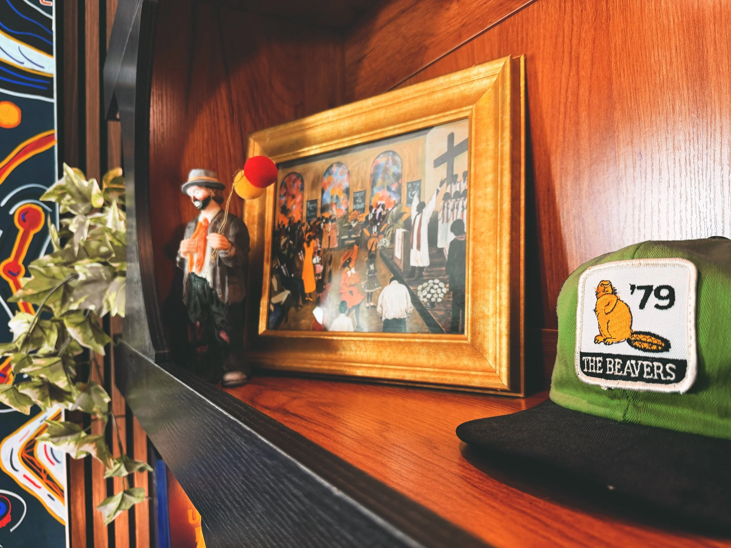 A framed painting of people gathering in a church interior hangs on a wooden wall. To the left, part of a pin art or graffiti art piece is visible. A decorative balloon figure is positioned in front of the painting, and a green patch with a lion and the quote 'The Beavers' is seen at the bottom right corner.